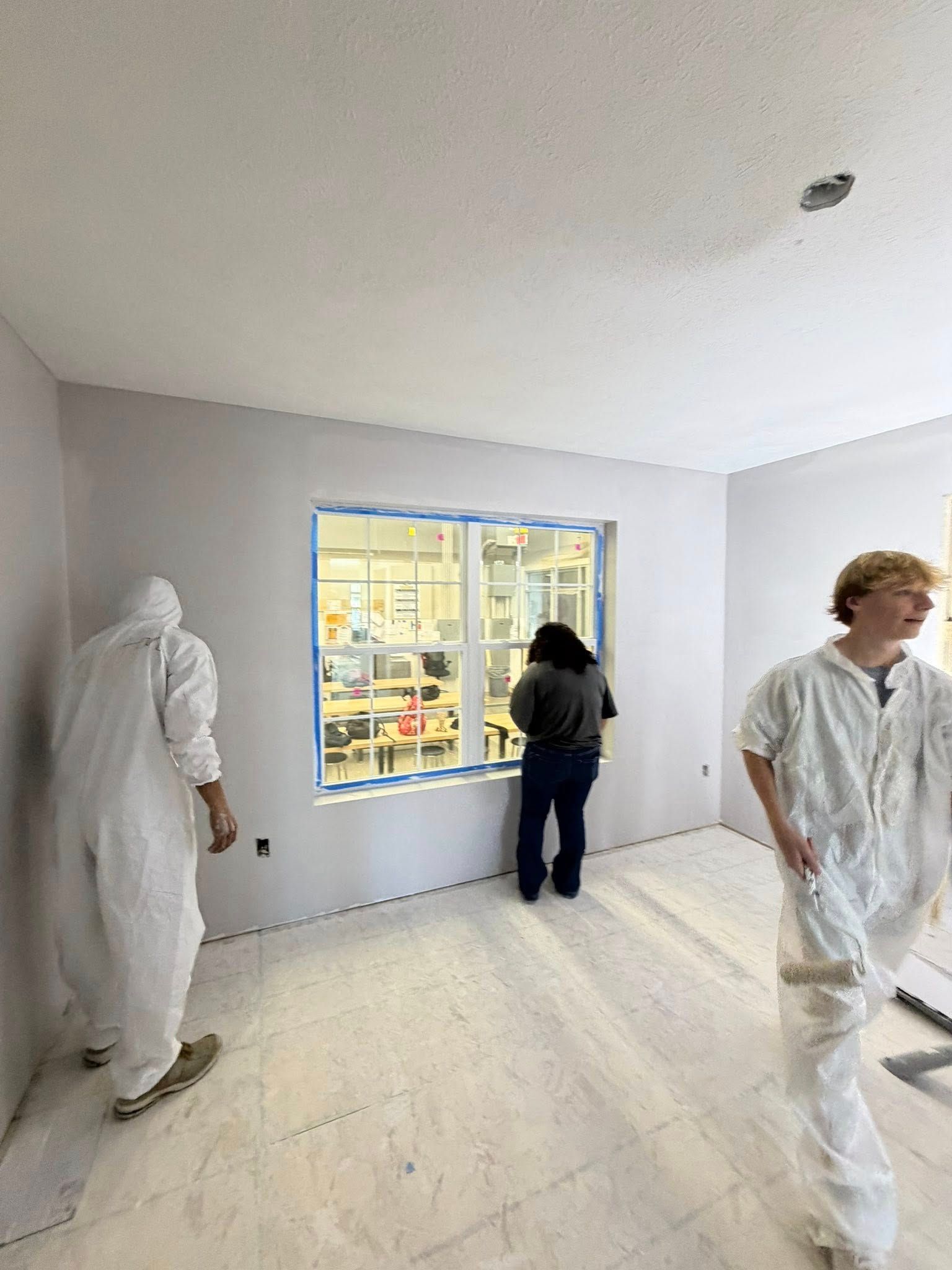Two people in protective suits and one in casual clothing work in a room with light gray walls and a taped-off window.
