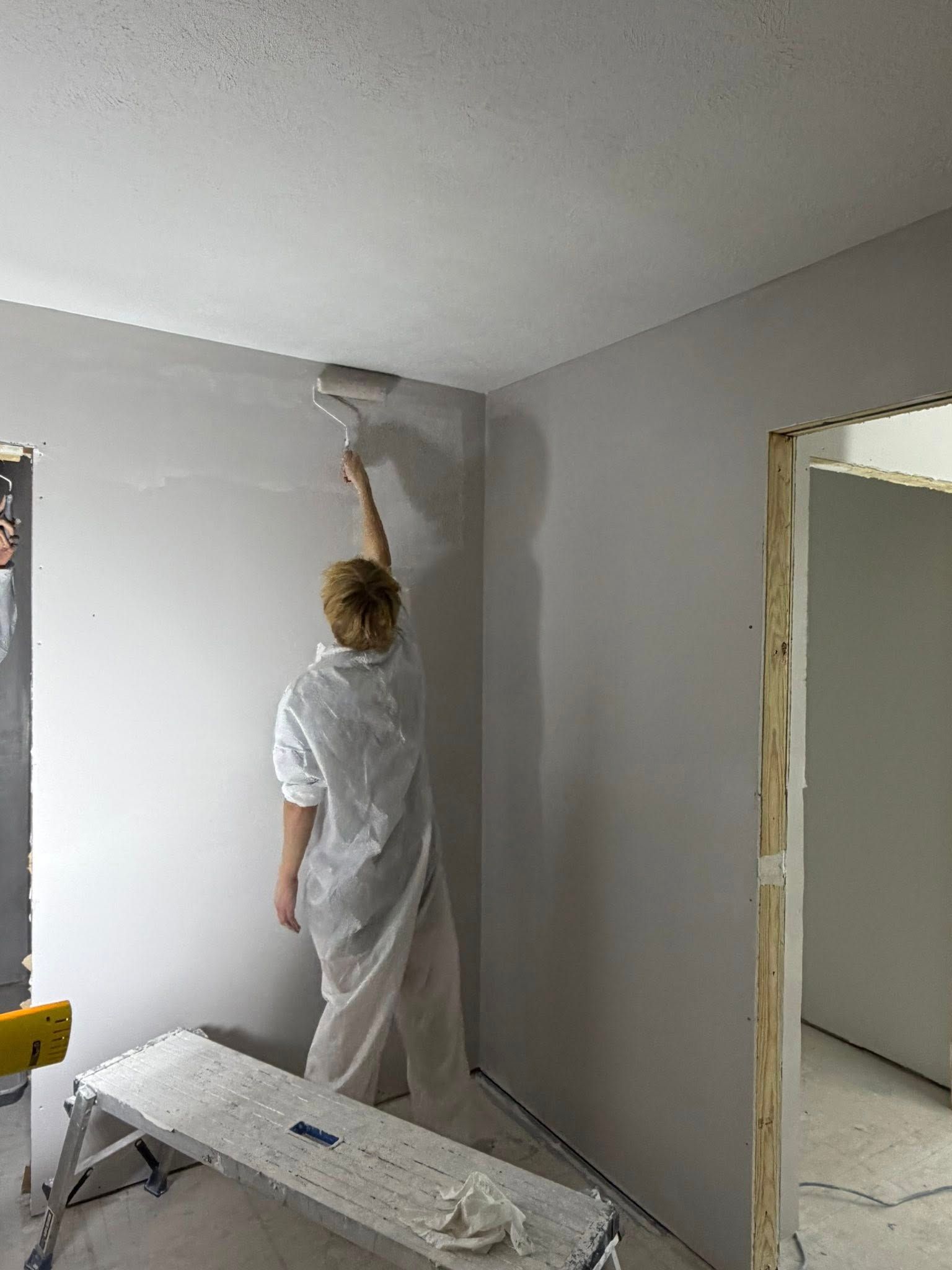 A person wearing white coveralls uses a paint roller to apply gray paint to a wall in an unfinished room.