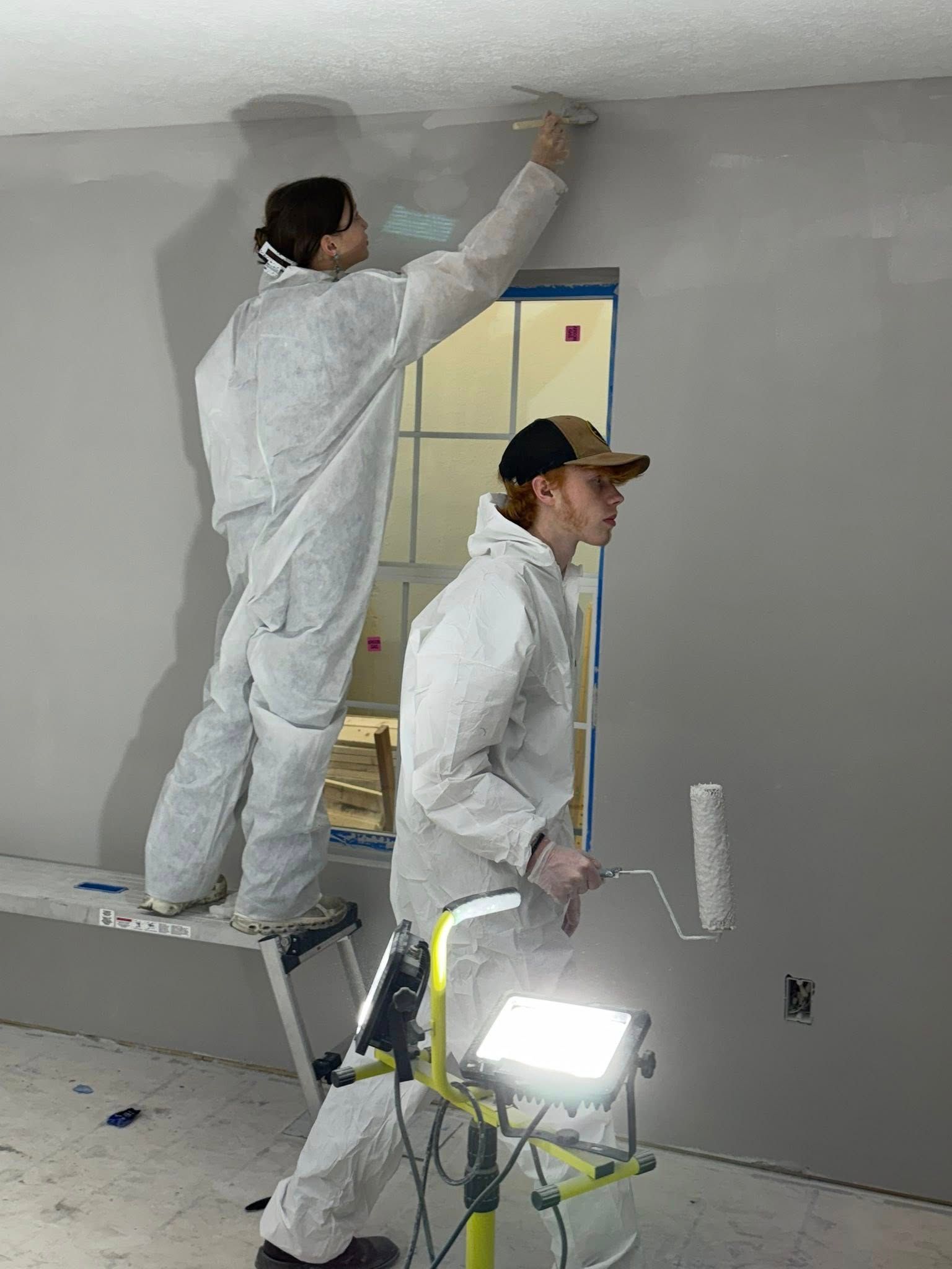 Two people in white protective suits paint a wall gray, one standing on a ladder and the other holding a paint roller.