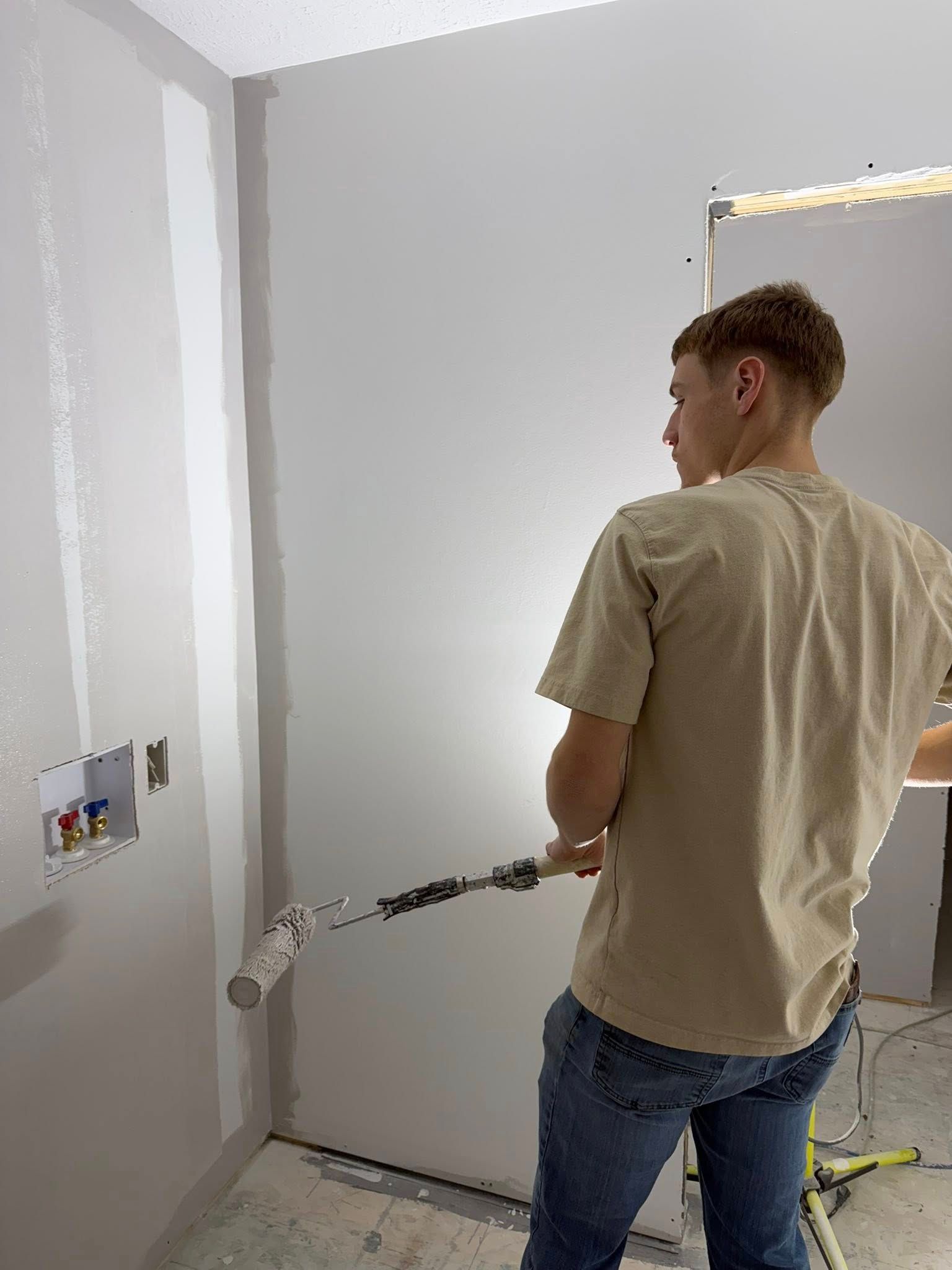 A person painting a white wall with a roller in a room under construction.