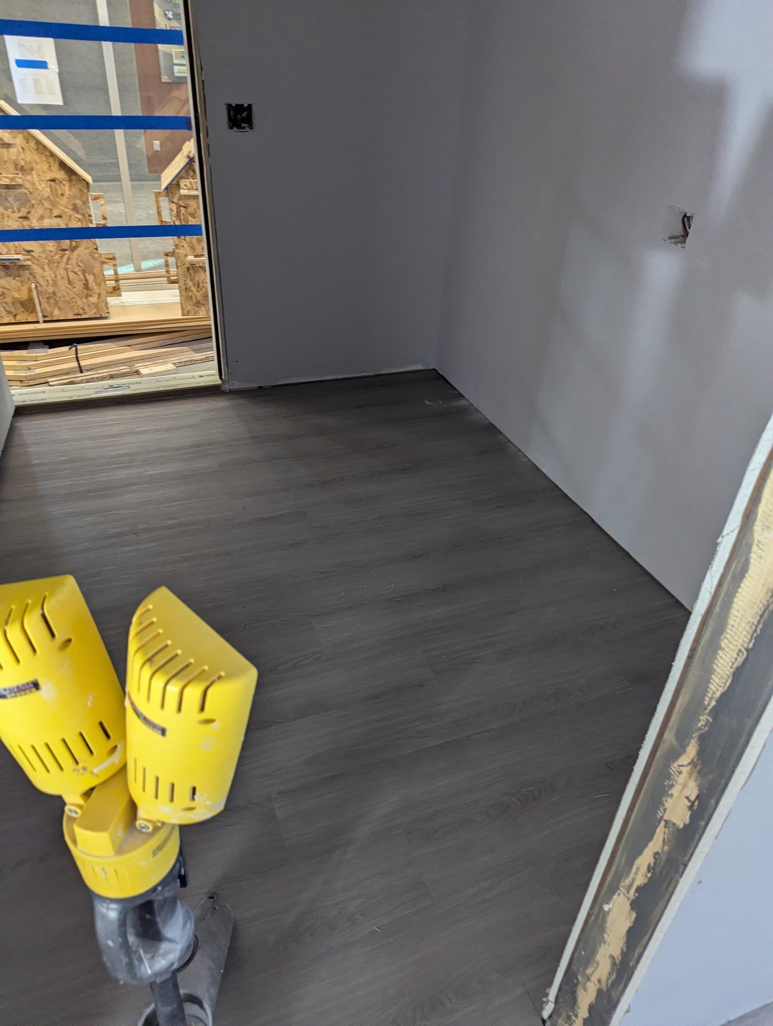 An interior room under construction with gray wood-look flooring and a yellow industrial work light in the foreground.