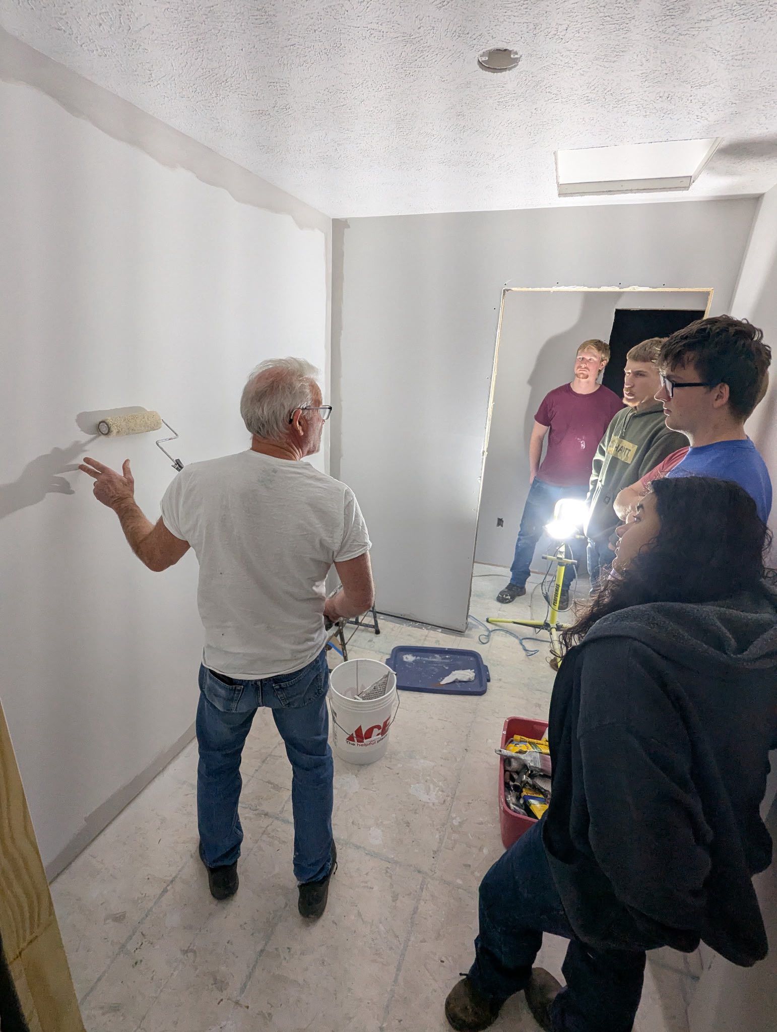 An instructor demonstrates wall painting techniques to a group of students in a room under renovation.
