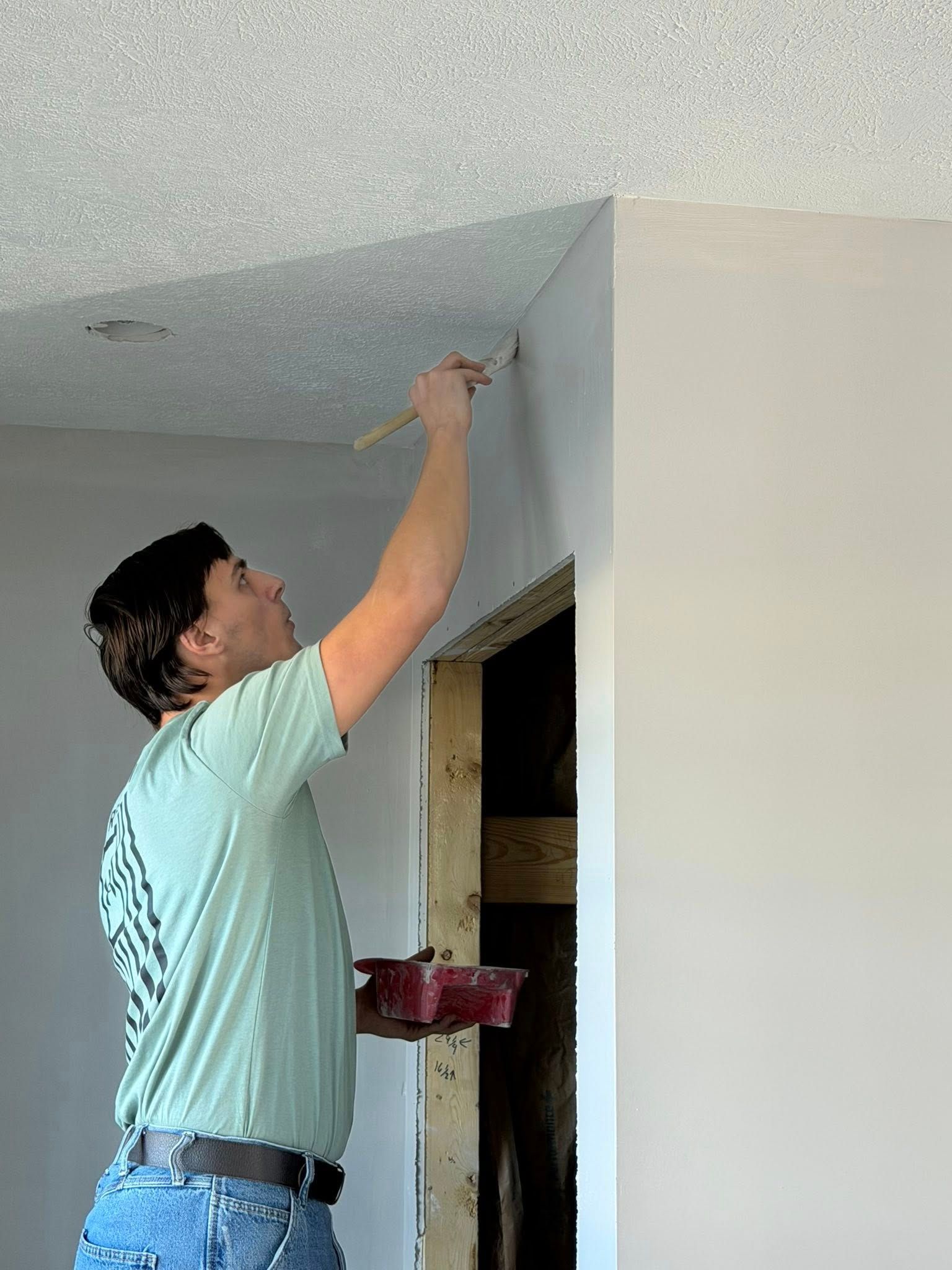 A person in a light green shirt uses a brush to paint the corner where a textured ceiling meets a wall.