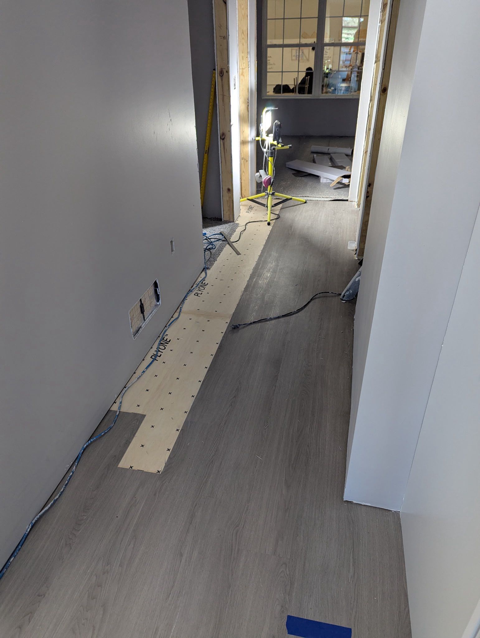 An interior hallway under construction, featuring light-colored flooring, exposed wall edges, and a portable work light.