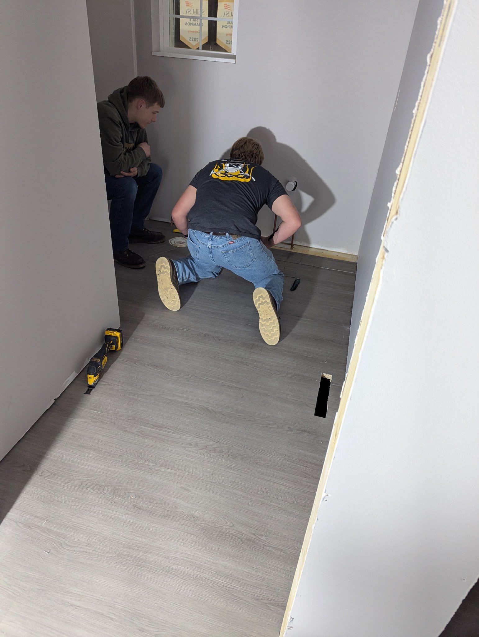Two people working on a room's grey subfloor, with one kneeling near a wall and another crouching nearby.