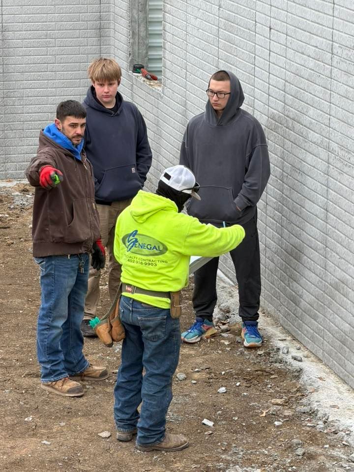 Four people stand in a dirt lot by a grey brick wall. A person in a bright yellow hoodie points at a document.