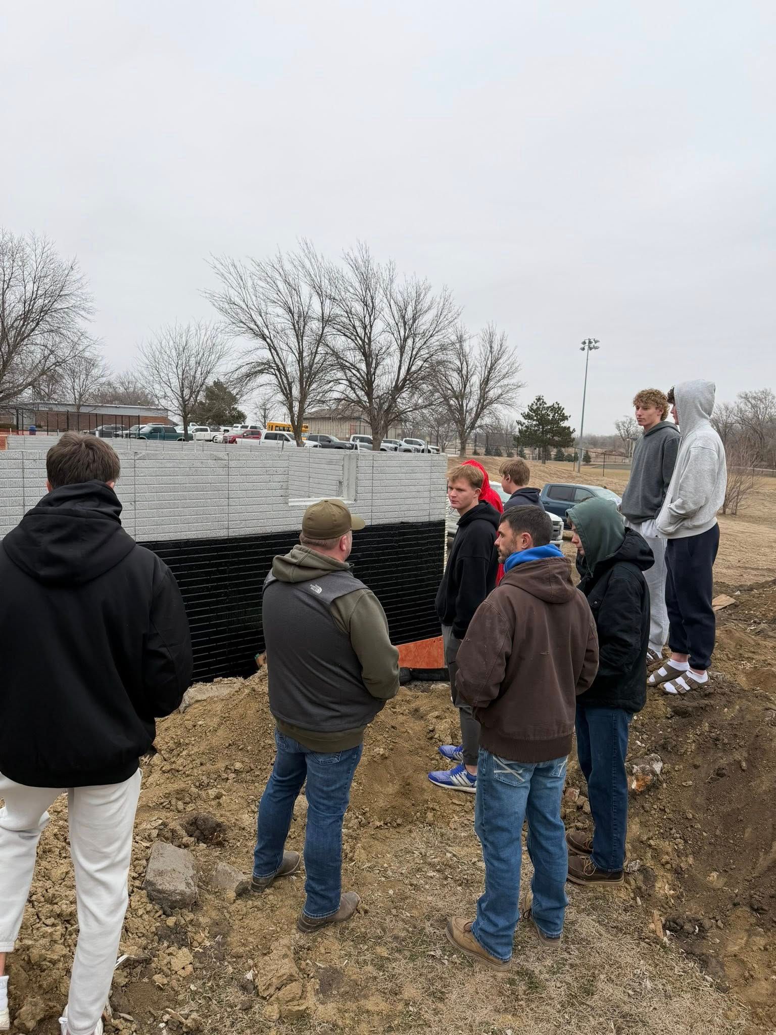 A group stands outdoors observing a section of black plastic grid material installed into a trench at a construction site.
