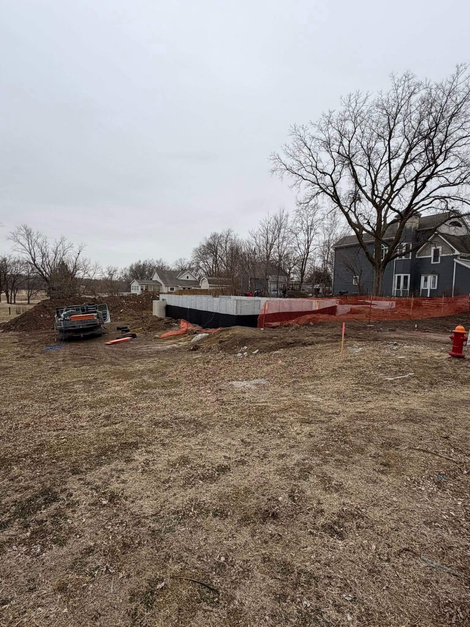 A construction site with a concrete foundation outline surrounded by orange safety fencing in a field with a house nearby.