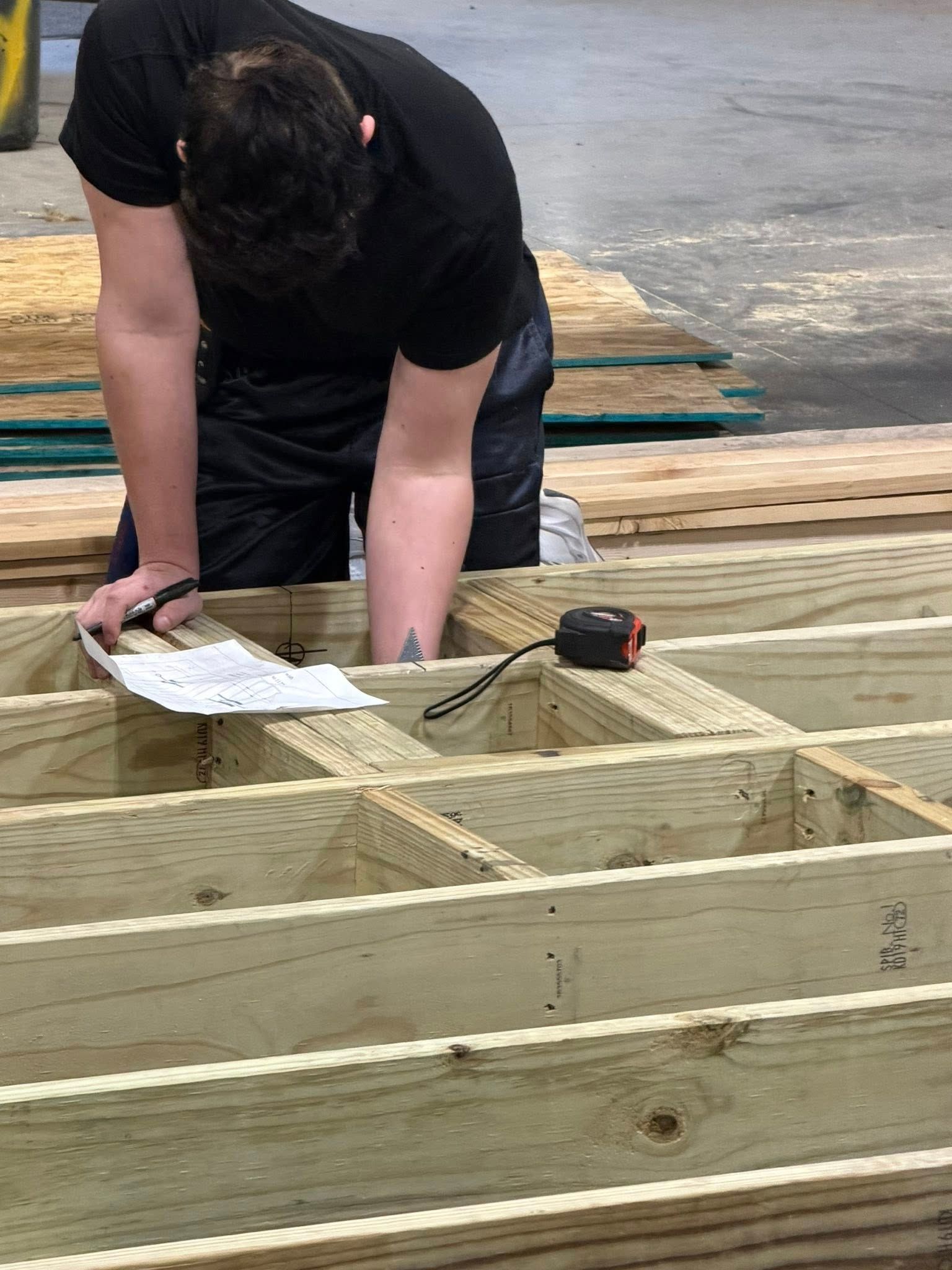 A person in a black shirt works on a wooden deck frame, marking dimensions on a paper with a tape measure nearby.