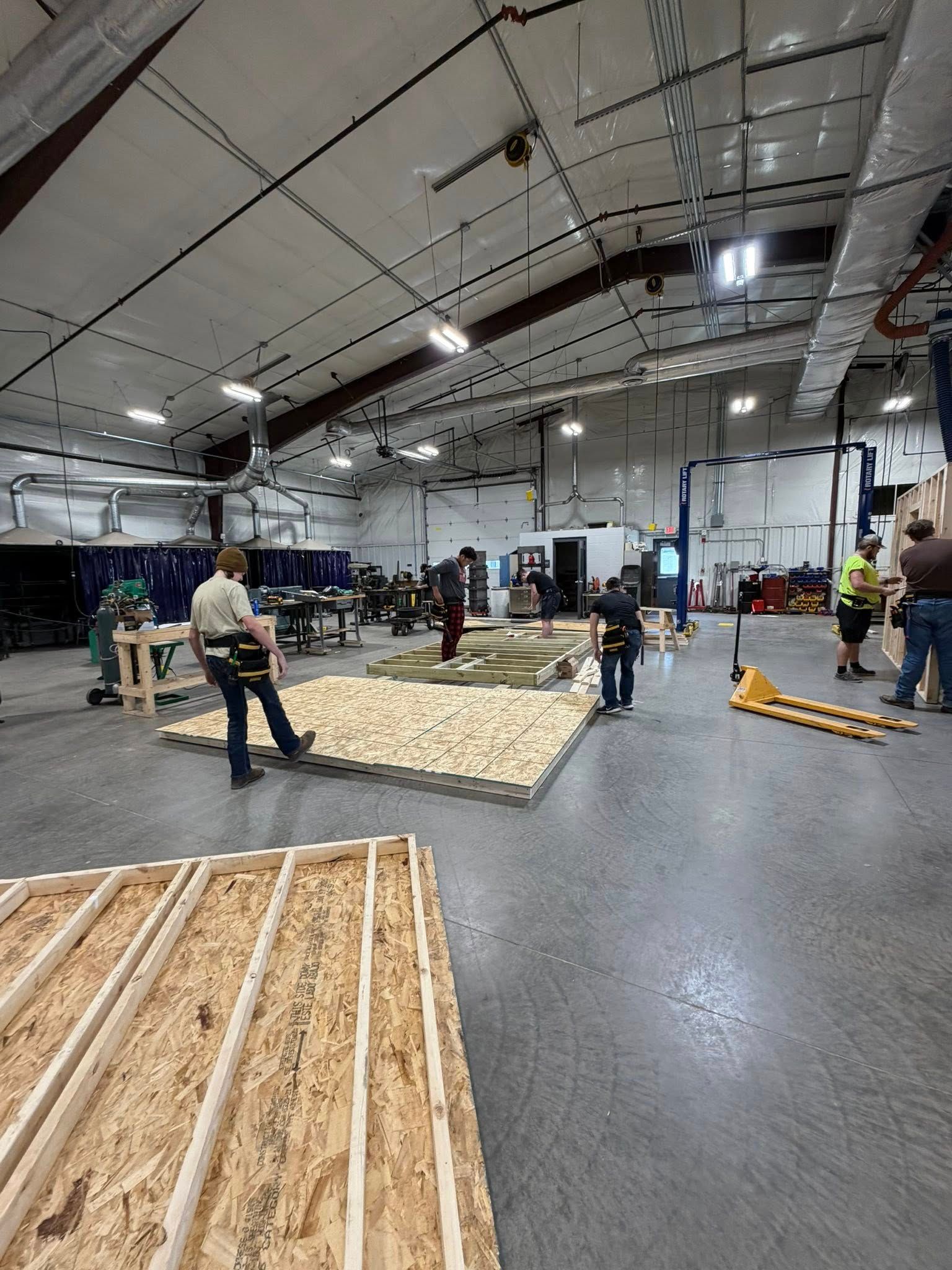 Workers assemble wooden wall frames in a large, bright industrial warehouse space.