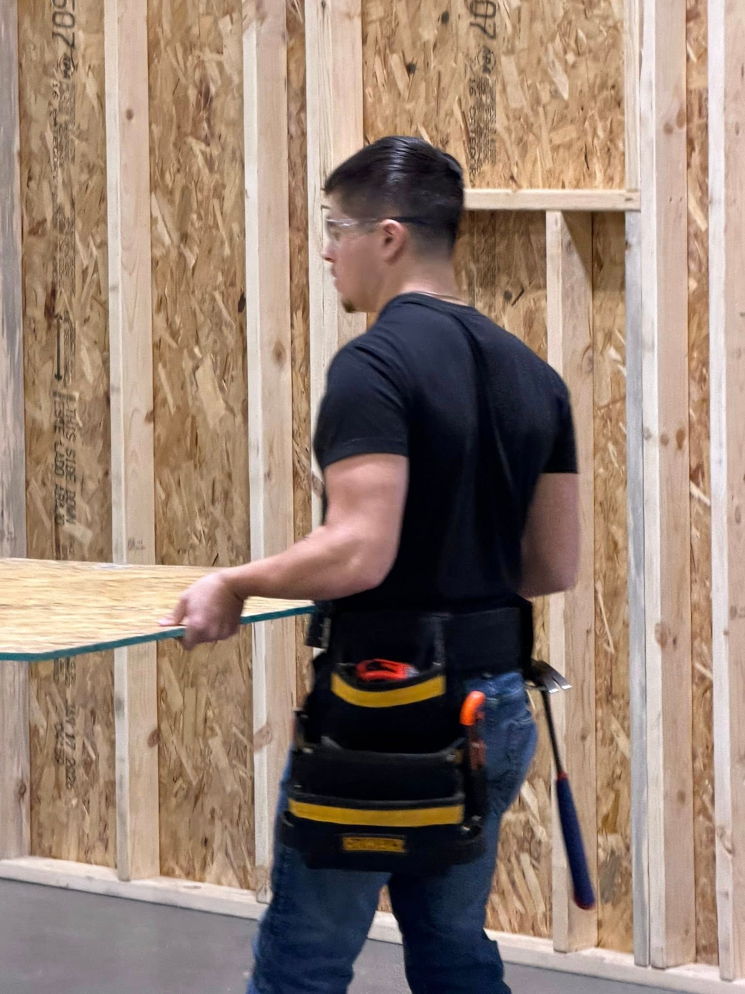 A person wearing a tool belt and safety glasses carries a wooden panel along a partially framed construction wall.