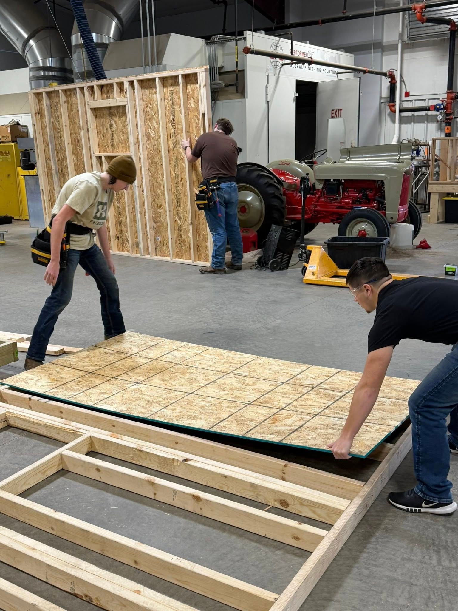 Two people working together to position a sheet of plywood onto a wooden floor frame in a workshop.