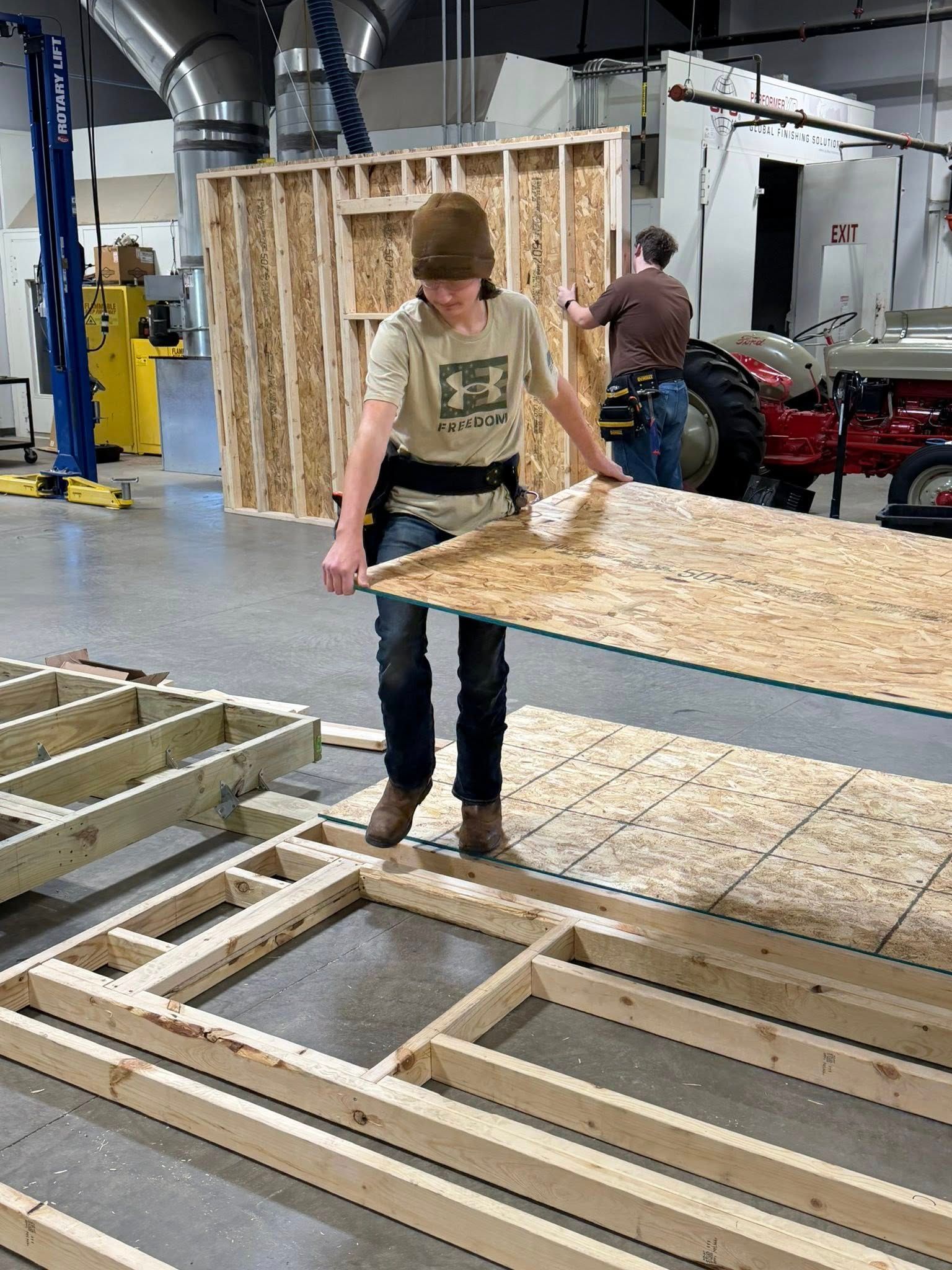 A person in a workshop wears a brown beanie and tan shirt while positioning a sheet of plywood over a wooden floor frame.