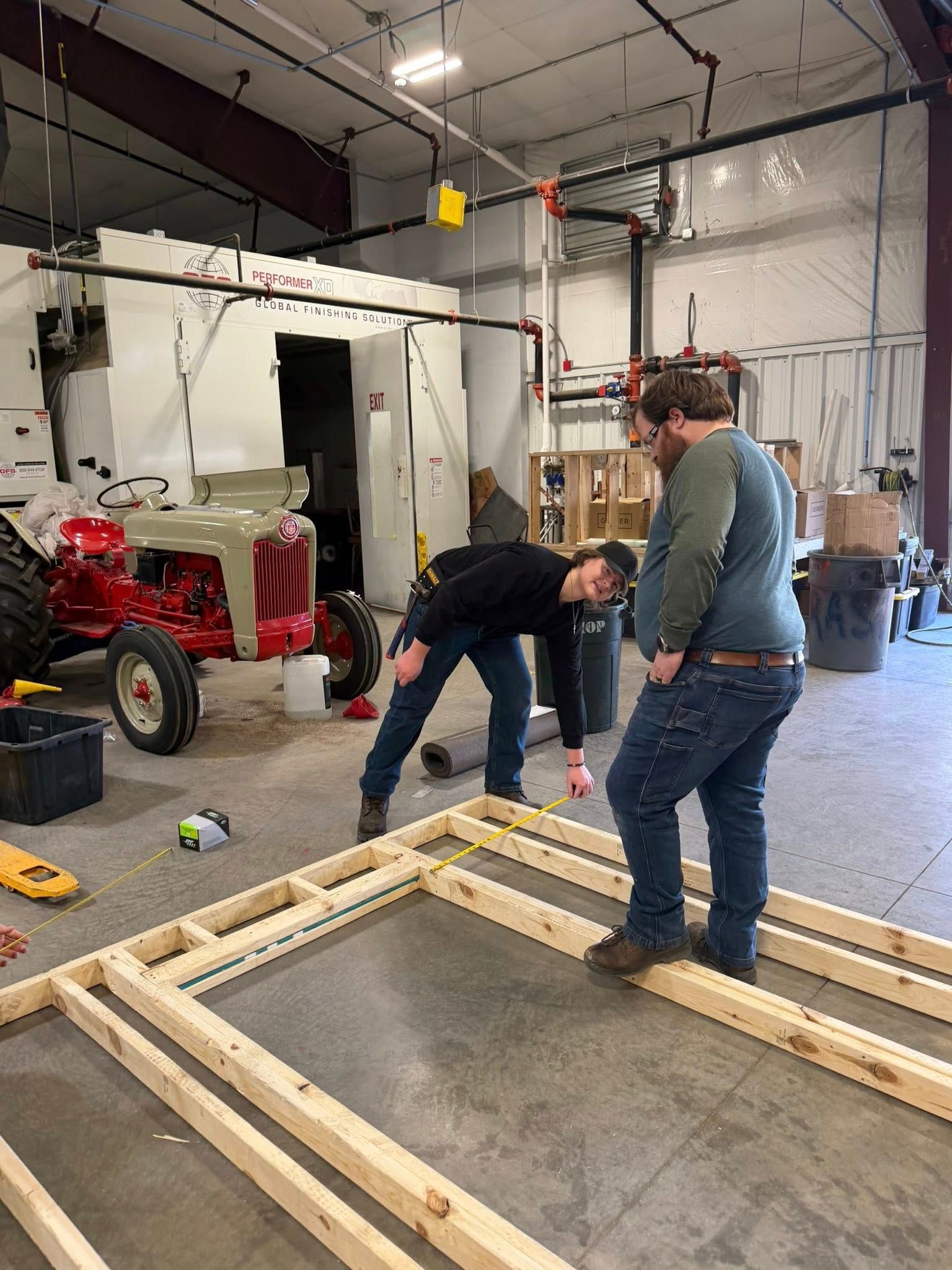 Two people working on a wooden wall frame in a workshop near a red vintage tractor.