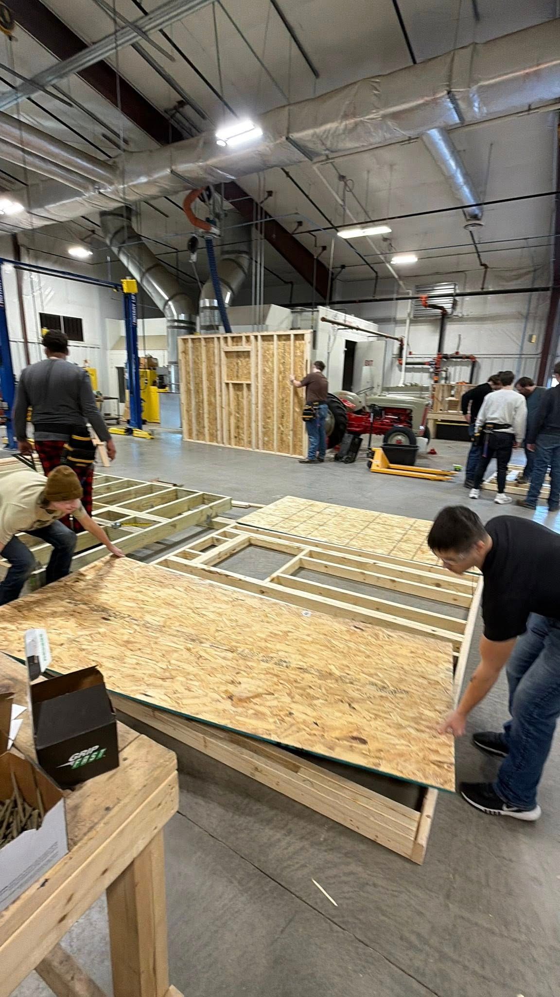 Students in a workshop setting assemble wooden wall frames and attach oriented strand board (OSB) sheathing.