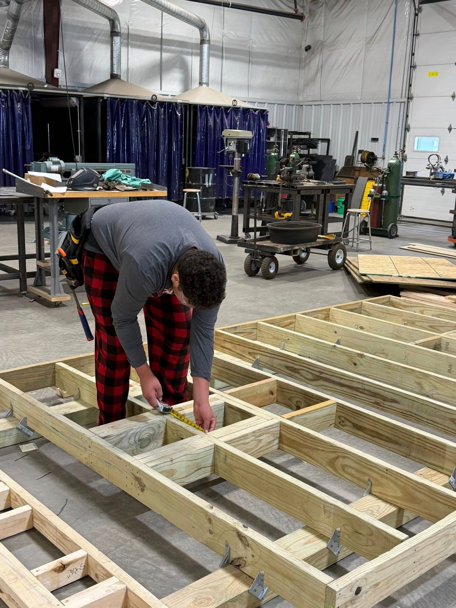 A person wearing plaid pants measures a wooden floor frame inside a large, industrial workshop with high ceilings.