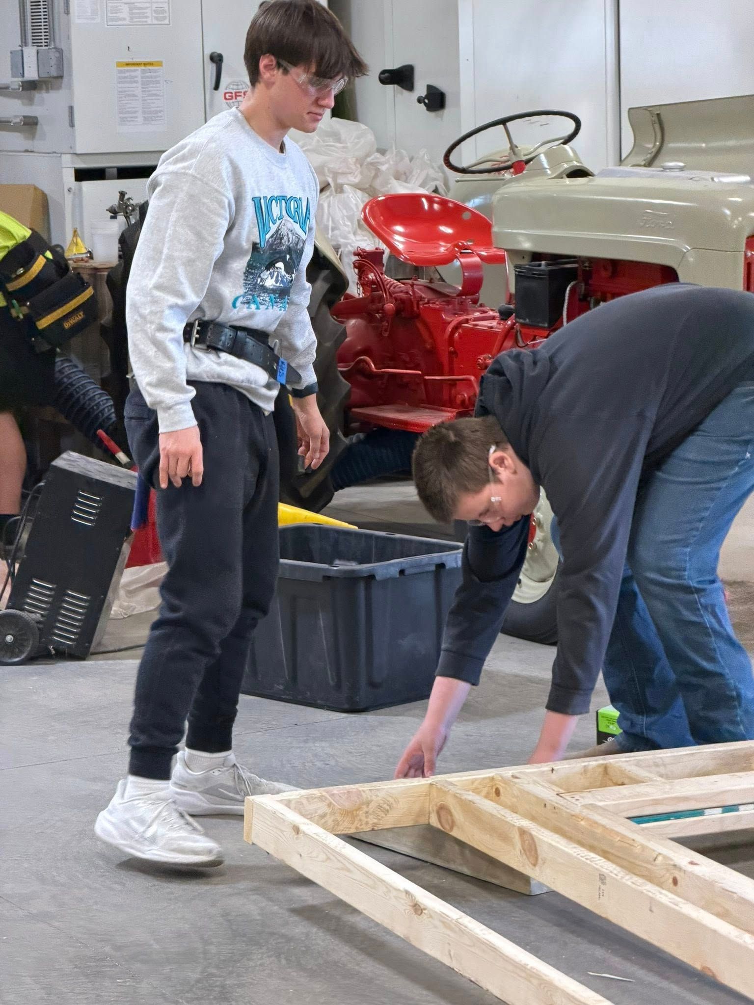 Two people work on a wooden frame structure in a workshop next to a red tractor.