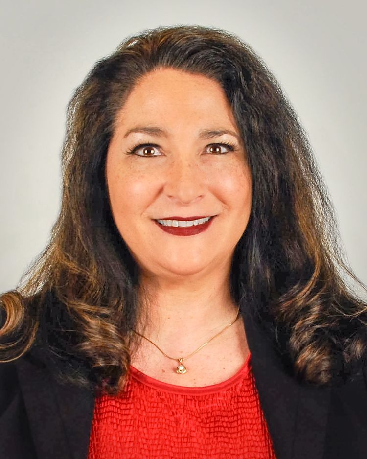A smiling person with long dark wavy hair, wearing a red textured top and a black blazer, poses against a gray background.