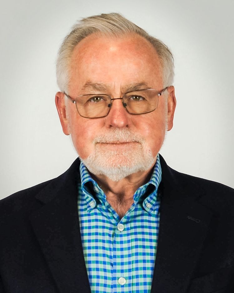 A person with gray hair and glasses, wearing a blue plaid shirt and a navy blazer, centered against a light background.