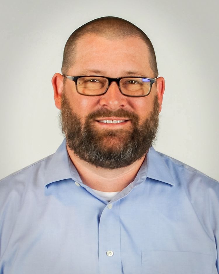 A person with a beard and glasses wearing a light blue button-up shirt against a plain, light gray background.