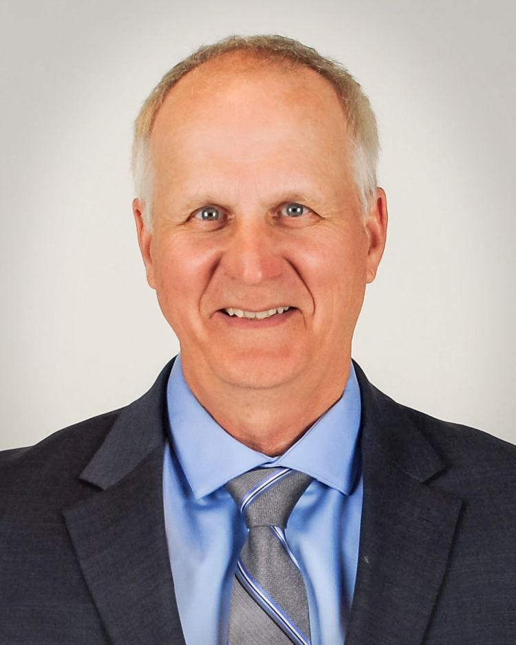 A person with short hair wearing a blue dress shirt, a striped tie, and a dark blazer against a plain gray background.