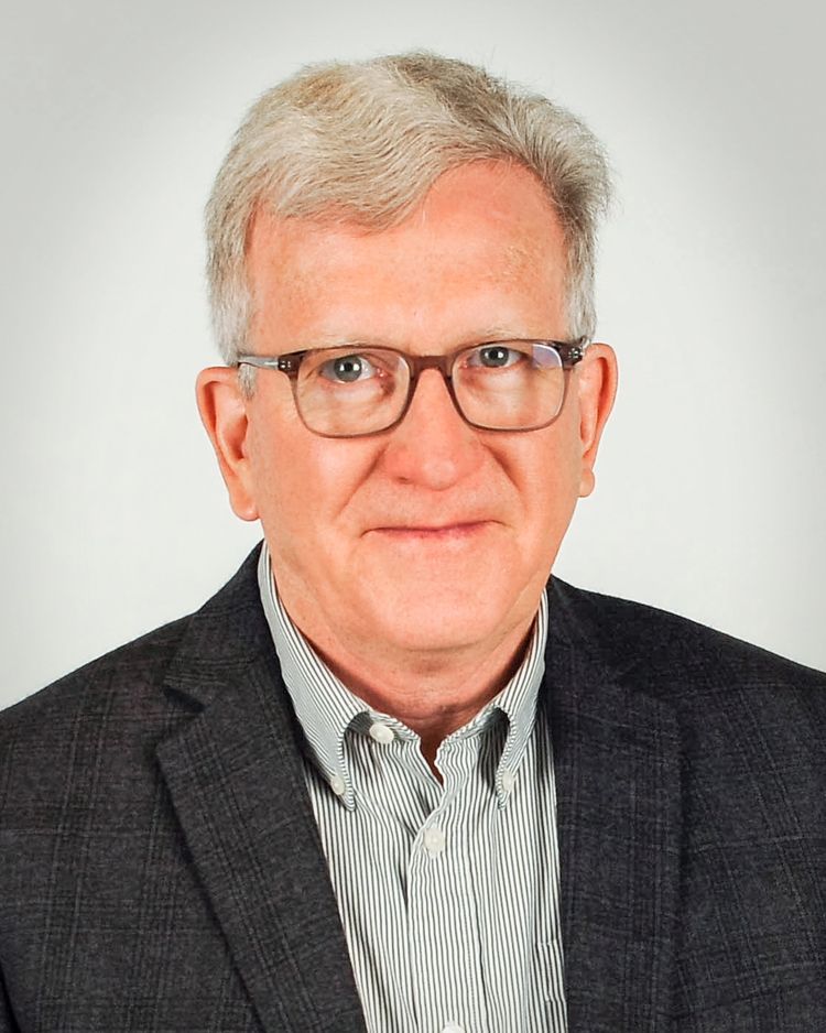 A headshot of a person with gray hair, wearing glasses, a striped collared shirt, and a dark blazer against a light wall.