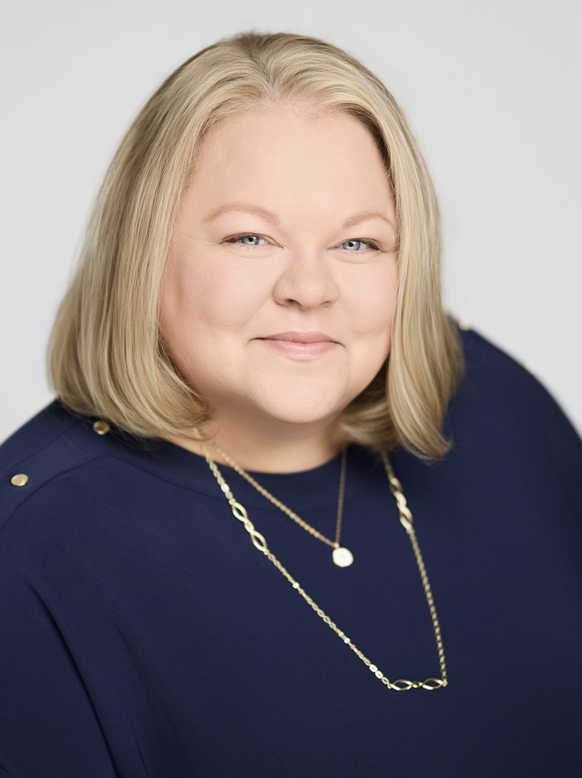Headshot of a person with blonde shoulder-length hair wearing a navy top and gold necklace against a light grey background.