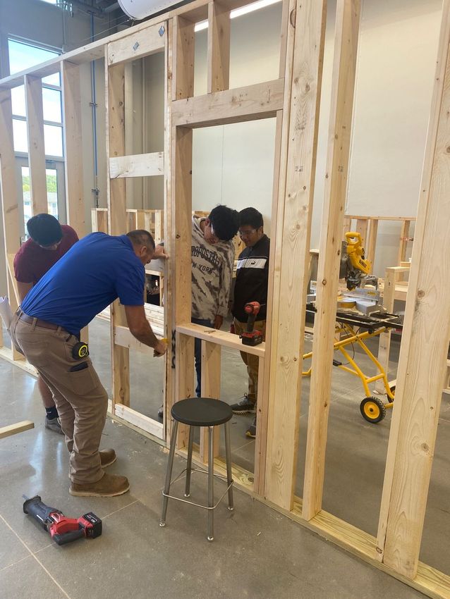 Three people construct a wood-framed wall in a workshop, with one person using a power tool on the frame.
