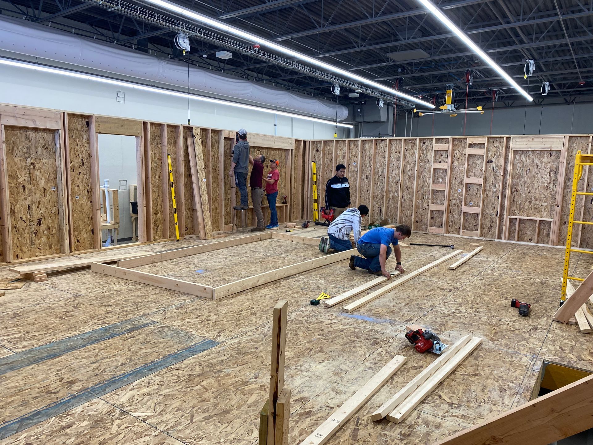 A group of people working together to construct the wooden frame walls of a house inside a large workshop.