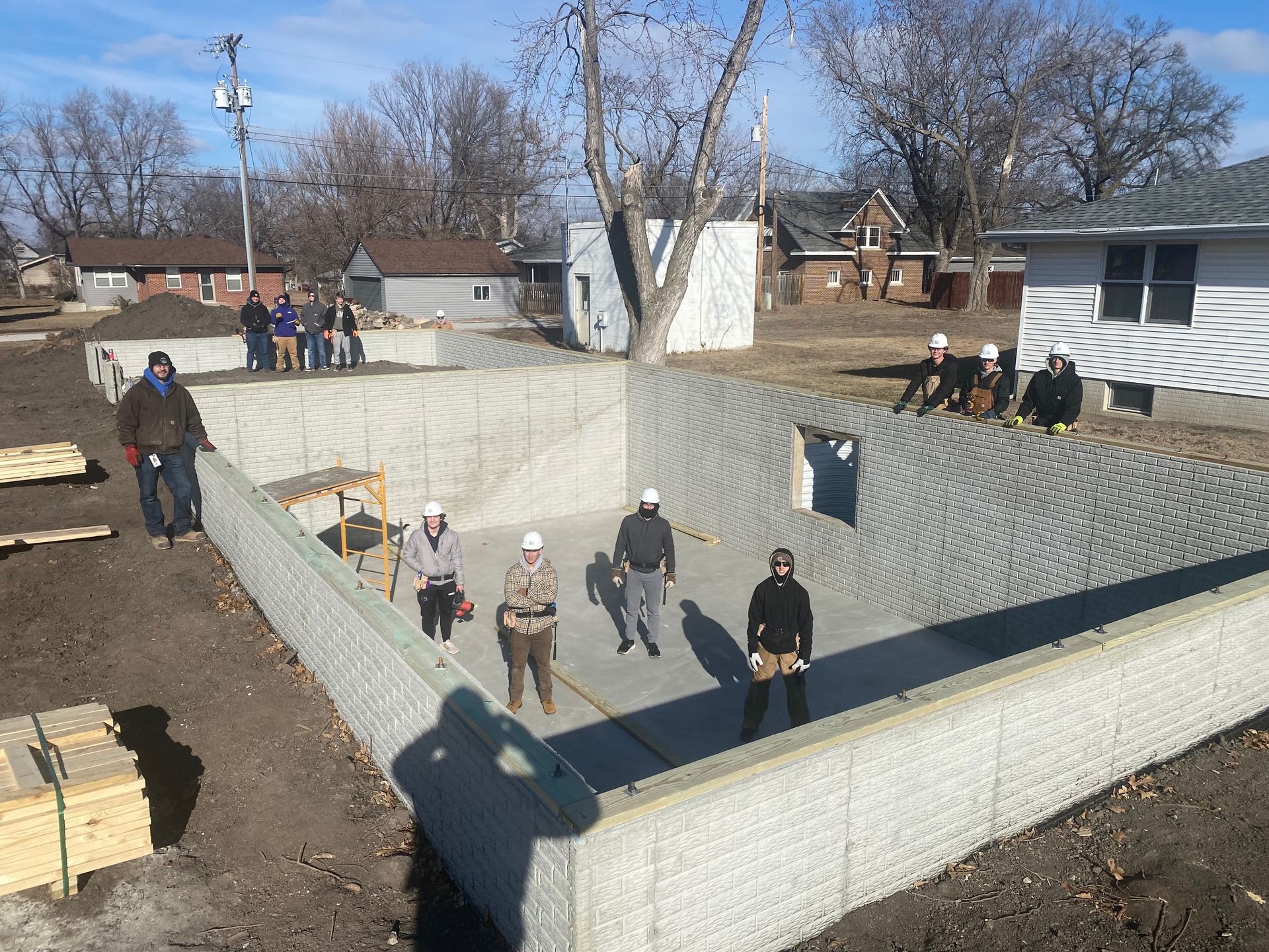 A group of workers in hard hats stands inside a concrete building foundation at a construction site.