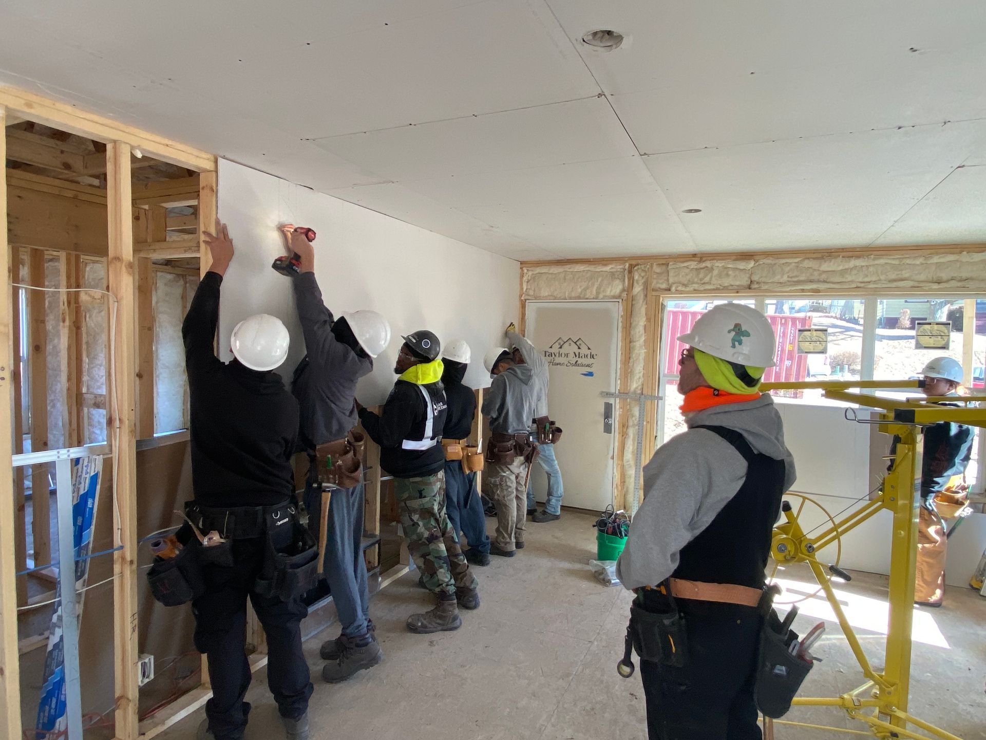 Construction workers in hard hats install drywall panels on interior wall framing in a residential building site.