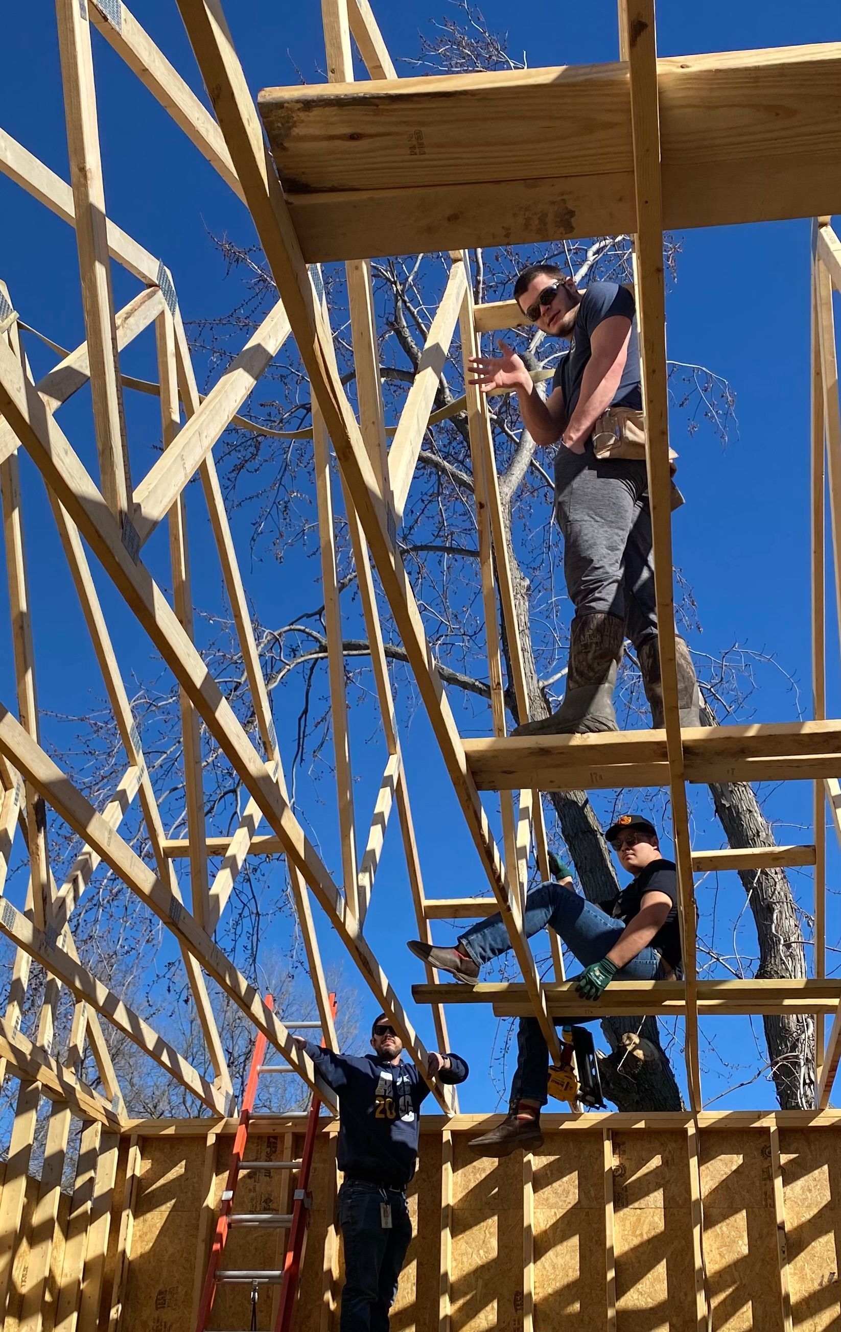 Three people work on the wooden framing of a building under a bright blue sky.