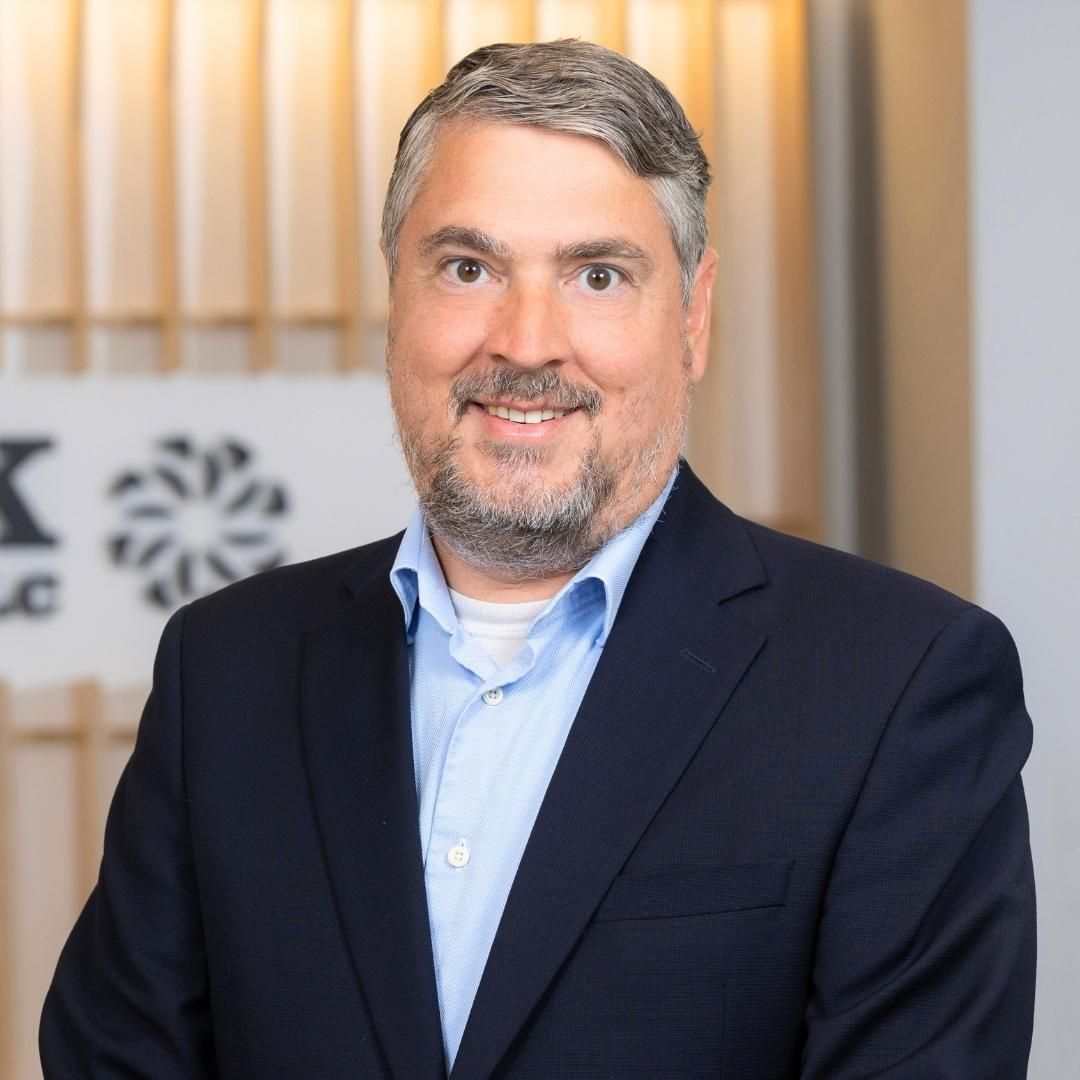 A professional headshot of a smiling man with a beard, wearing a blue button-down shirt and a navy blazer.