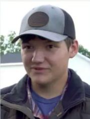 A person wearing a grey and black baseball cap with a brown circular patch, a plaid shirt, and a dark jacket.