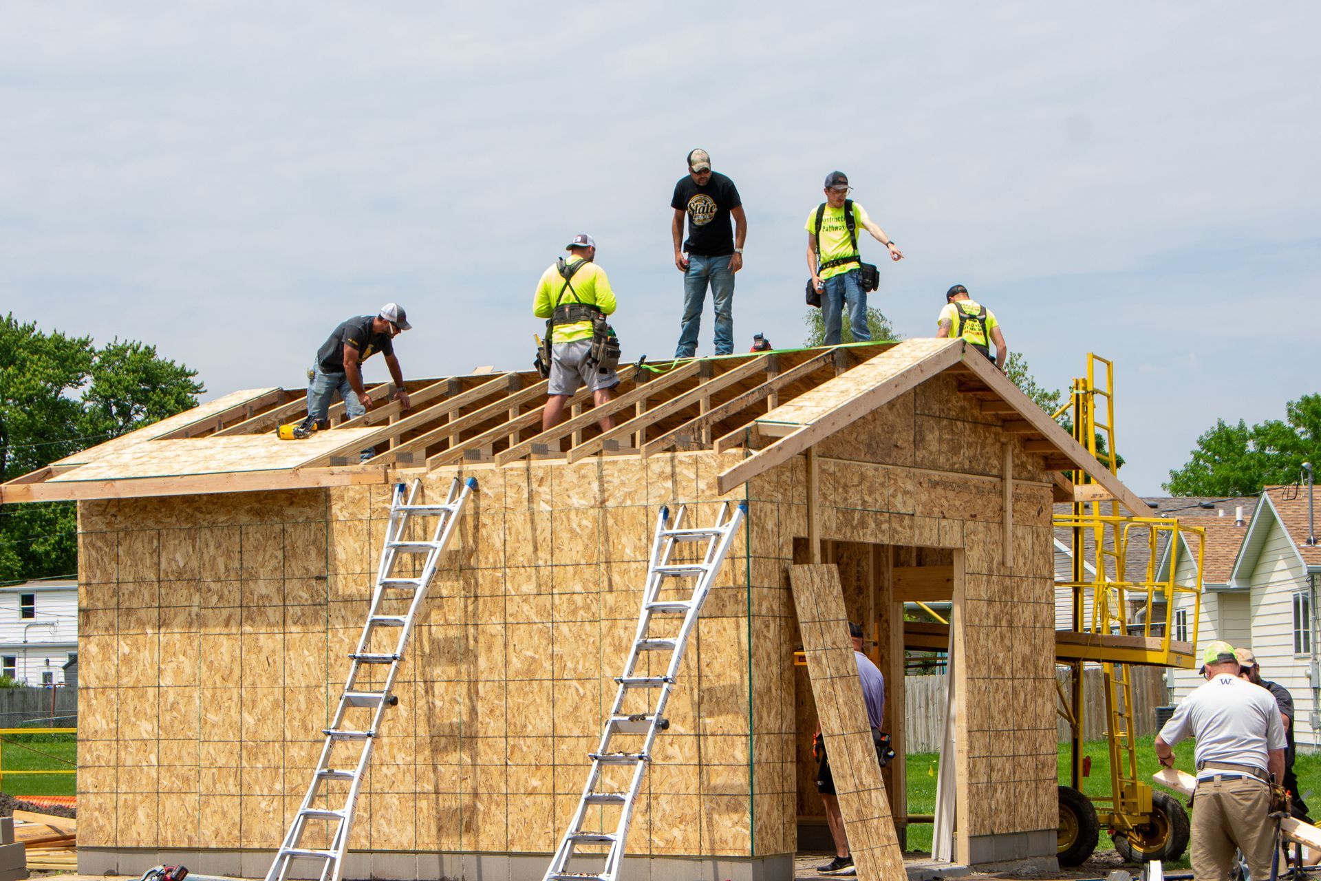Construction workers on the roof and ground of a wooden frame structure, using ladders and gear under a bright sky.