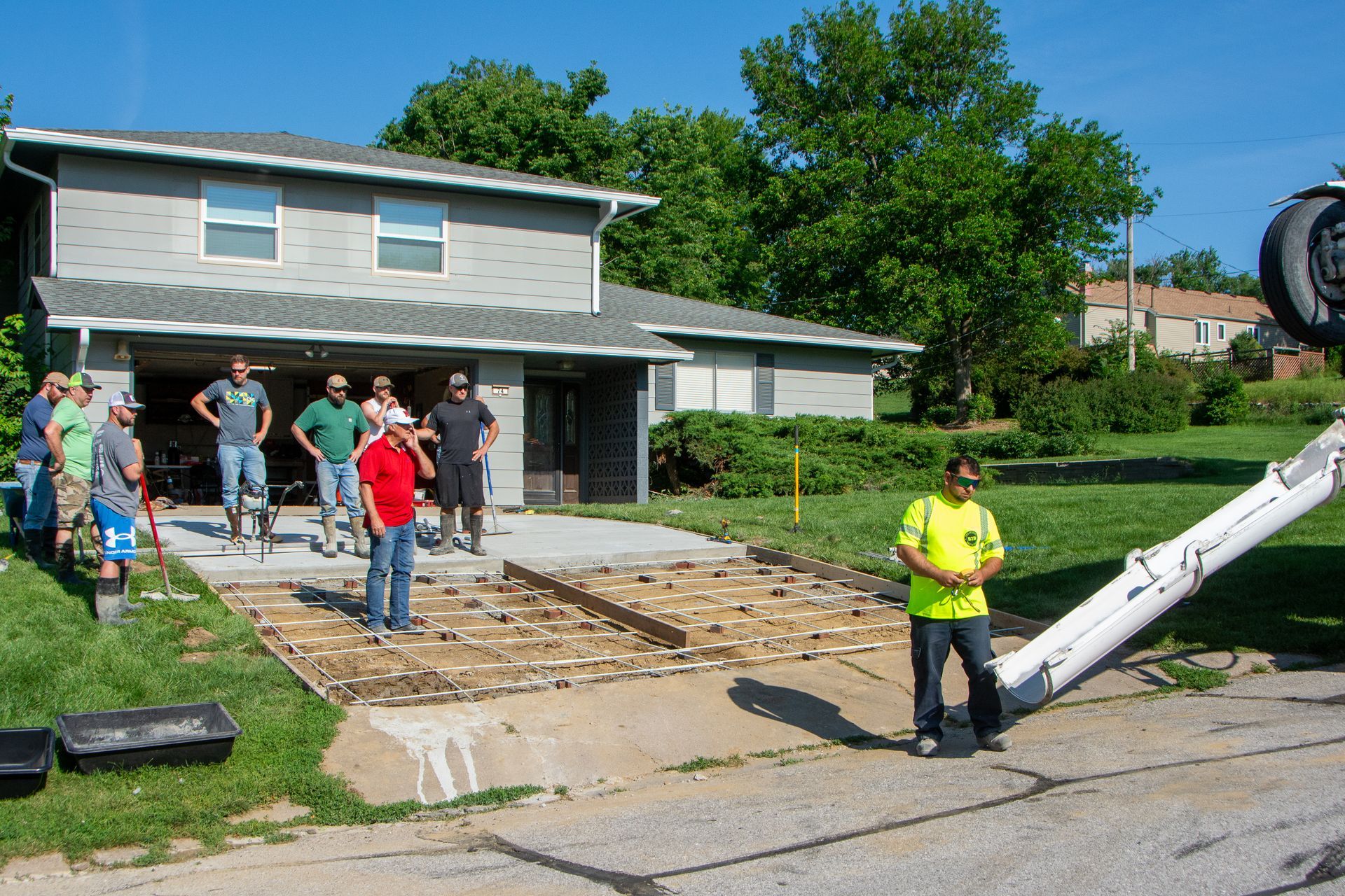 A construction crew stands by a concrete-poured driveway as a truck nozzle prepares to finish the job outside a house.