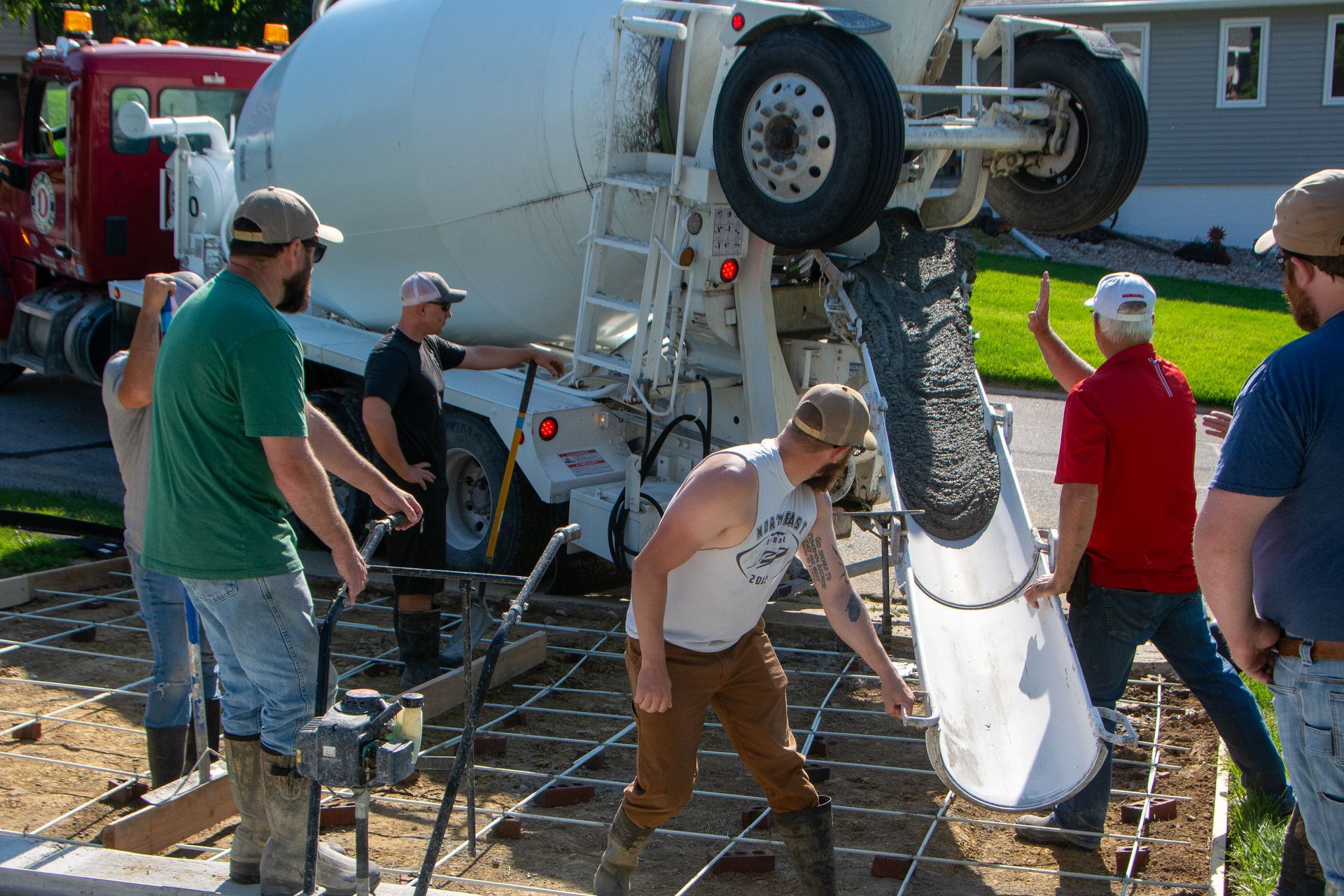 Construction workers pour concrete from a mixer truck into a wire-meshed frame in a residential yard.