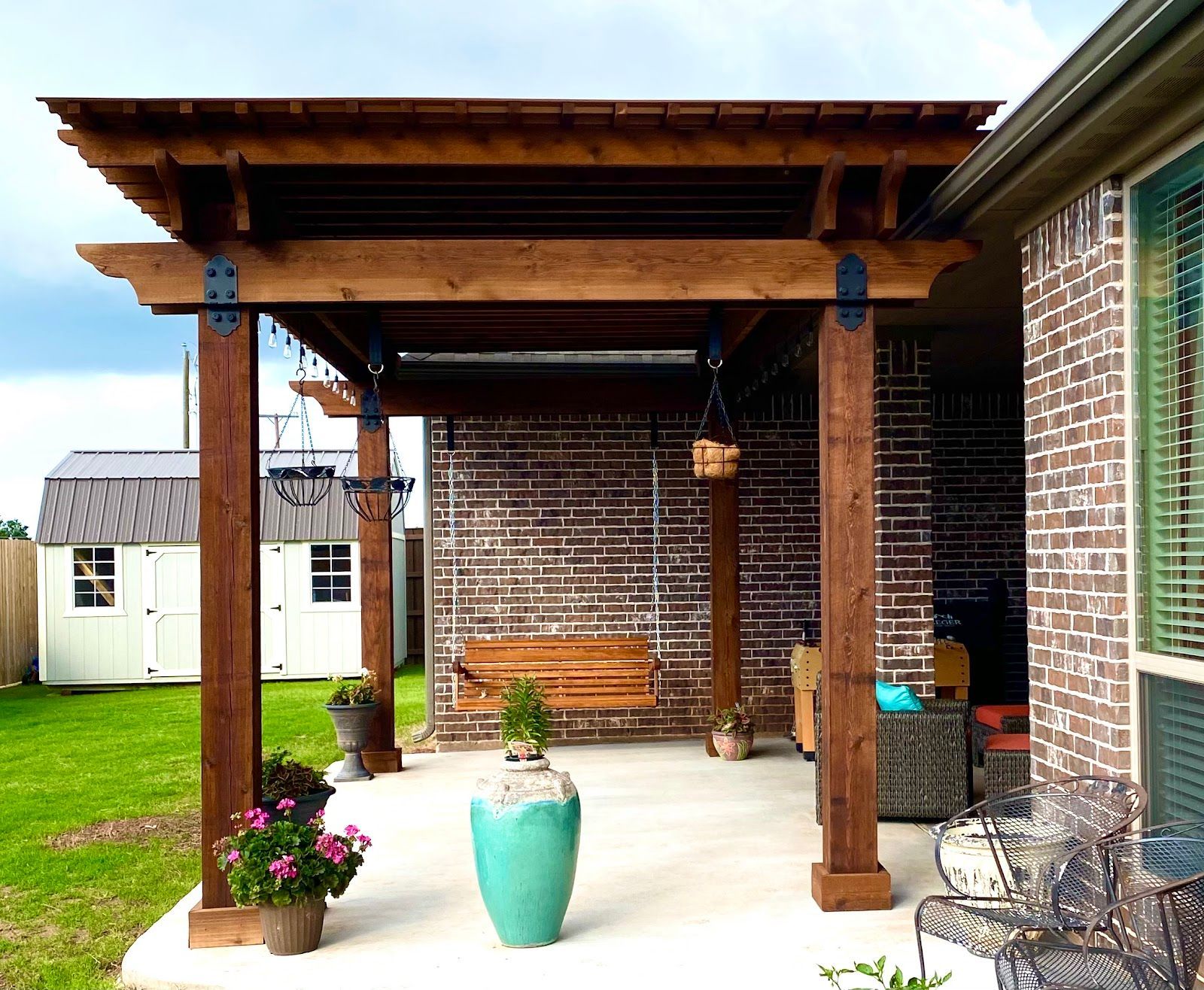 A wooden pergola with hanging swing on a concrete patio, beside a brick wall and house.