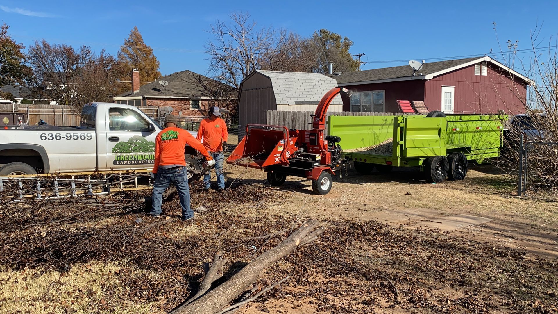 Three workers chipping wood debris on a lawn, with truck and trailer on a sunny day.