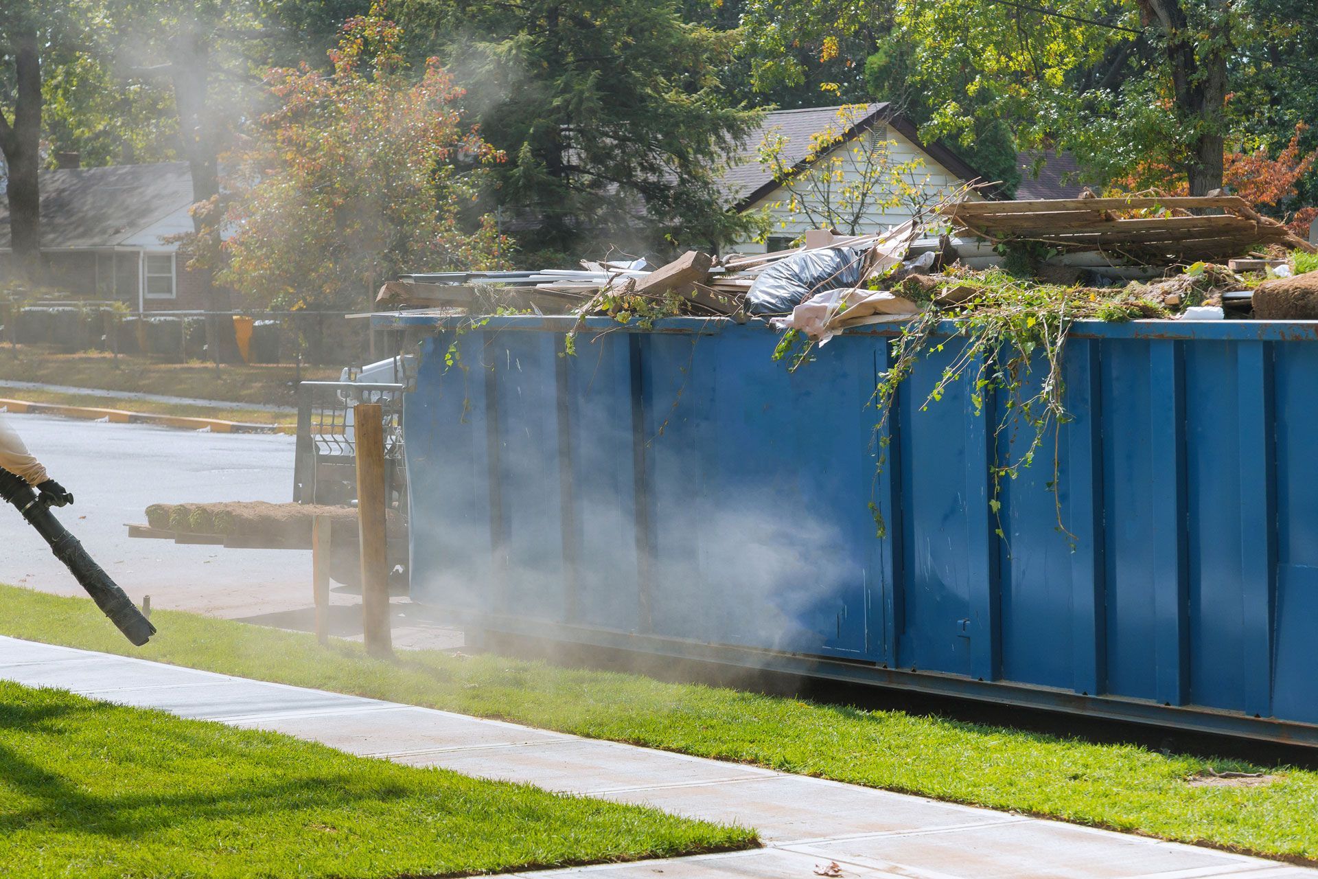 Blue construction dumpster smoking beside a sidewalk after a fire, with water spraying nearby