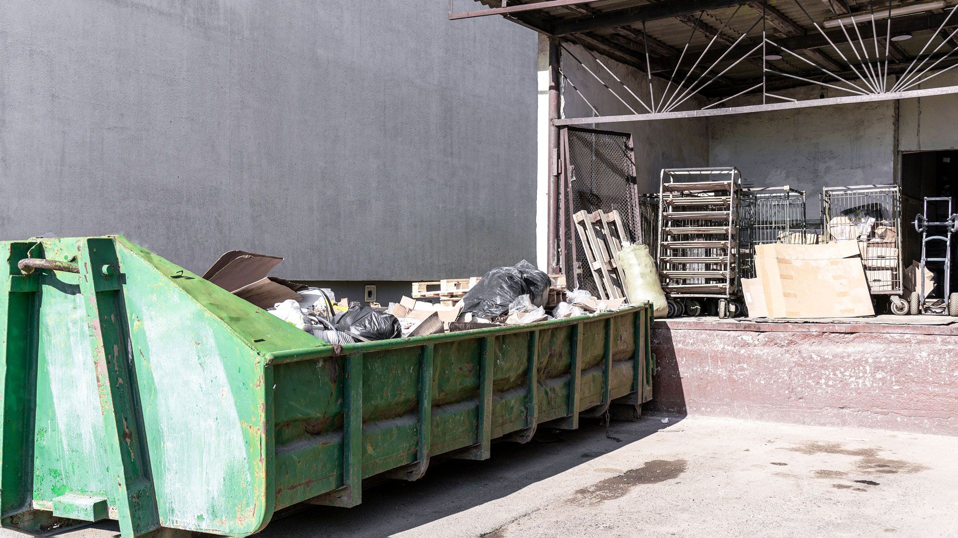 Green dumpster full of debris beside an open warehouse loading bay