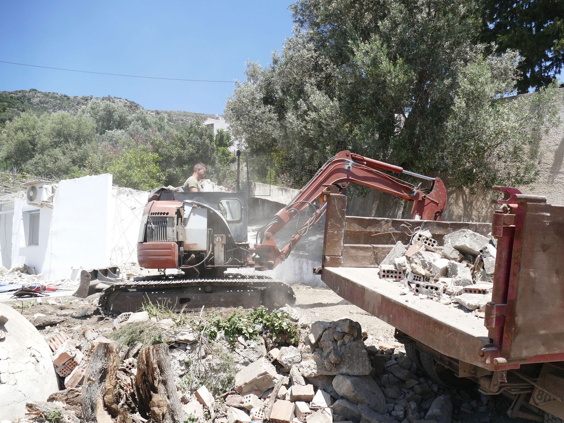 Excavator loading rubble into a red dump truck at a demolition site outdoors