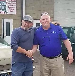Two men are shaking hands in front of a car.