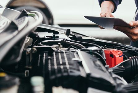 A man is looking under the hood of a car while holding a tablet.
