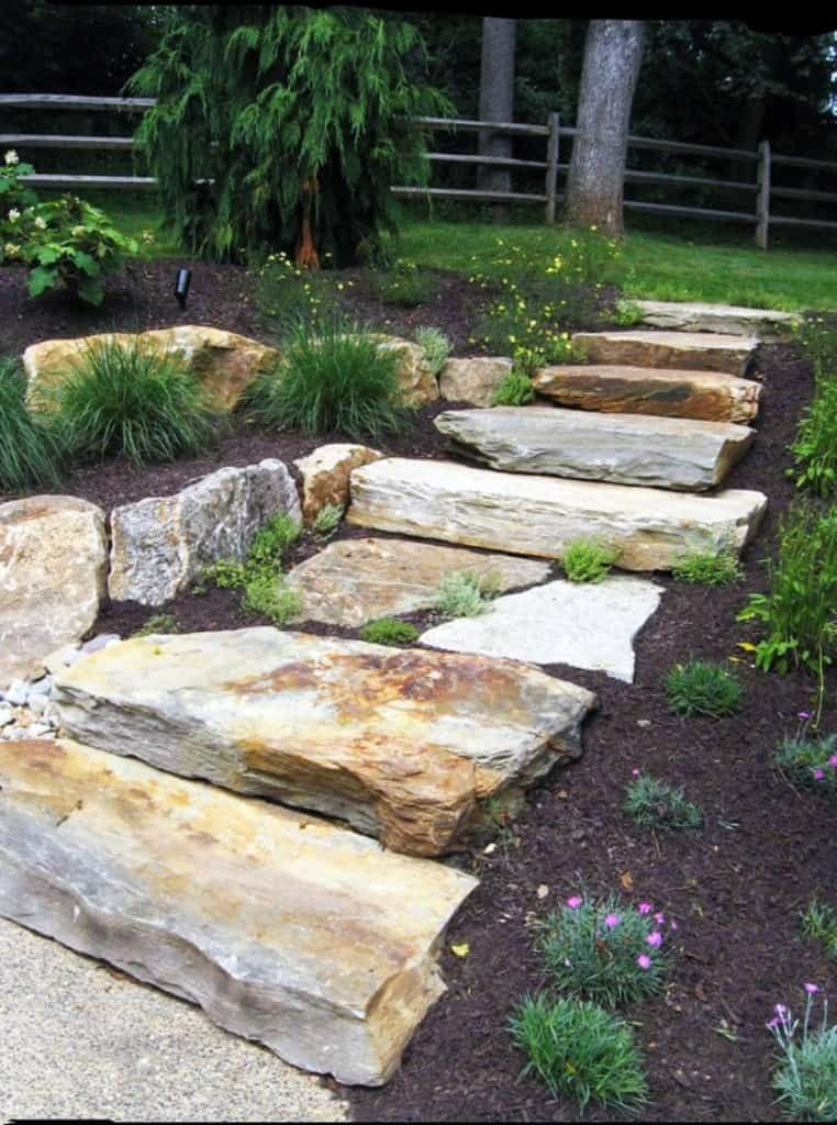 Stone steps ascend through a garden with mulch and various plants, leading toward a wooden fence.