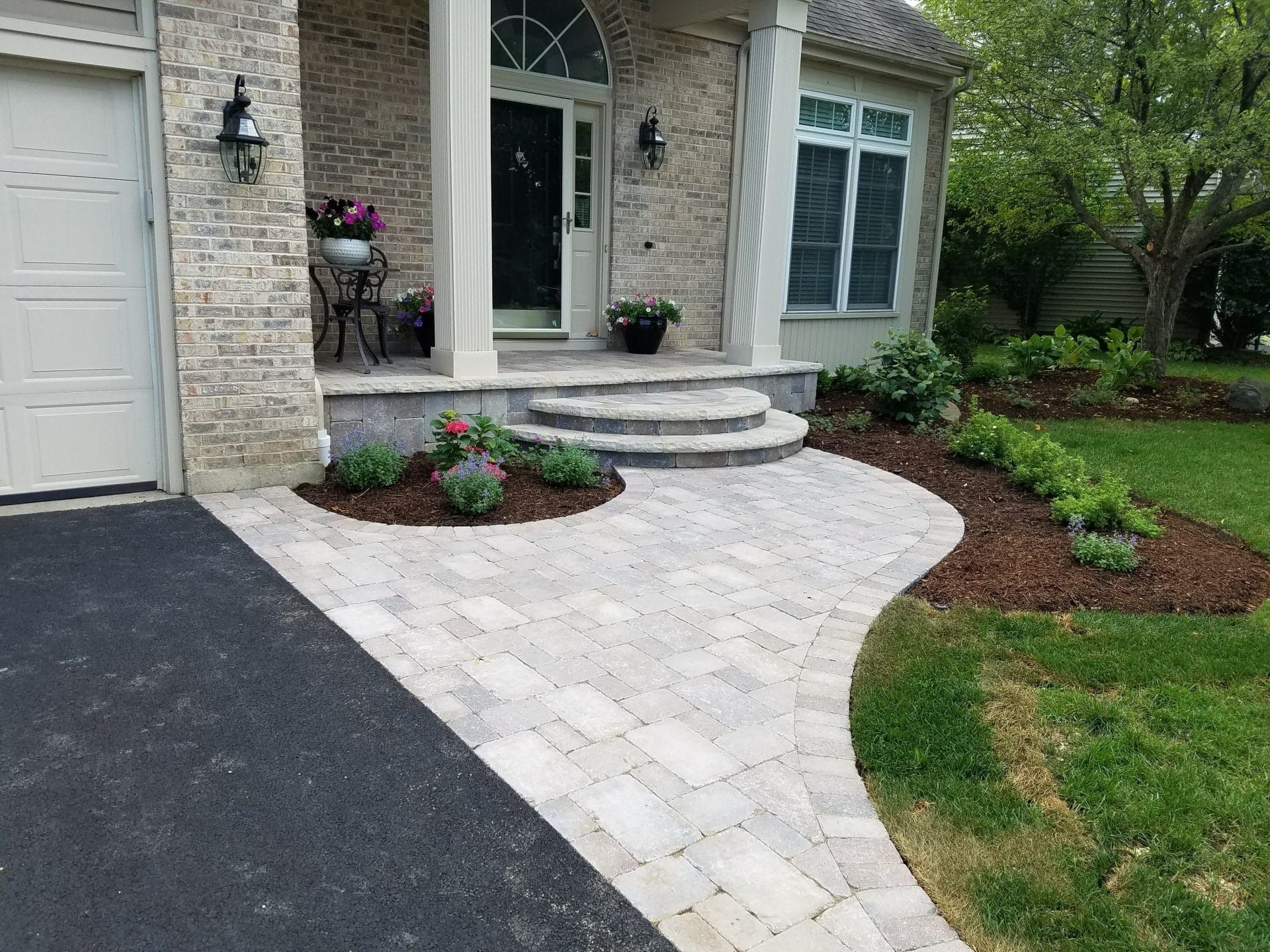 Brick walkway leading to a house's entrance with landscaping, steps, and a driveway.