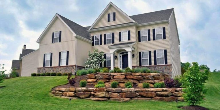Two-story beige house with black shutters, built into a grassy hill with stone retaining walls.