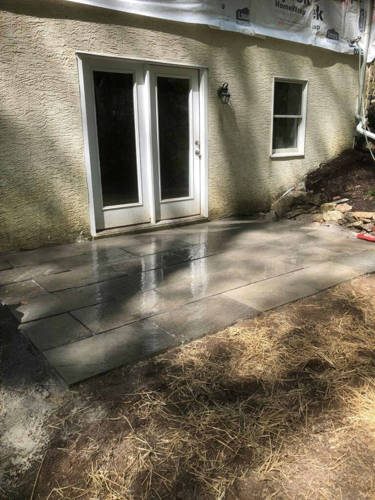 Exterior patio with wet, gray stone tiles, next to a building with double doors and a window.