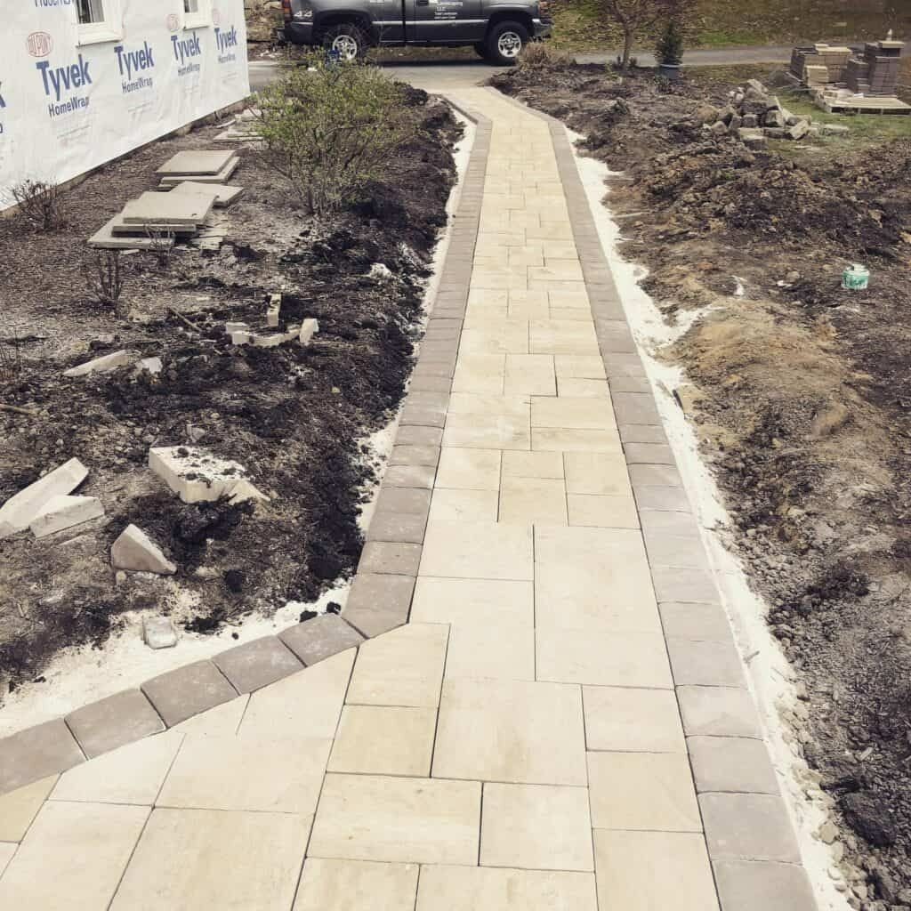 A newly constructed brick pathway with a dark border, next to a house and construction site.