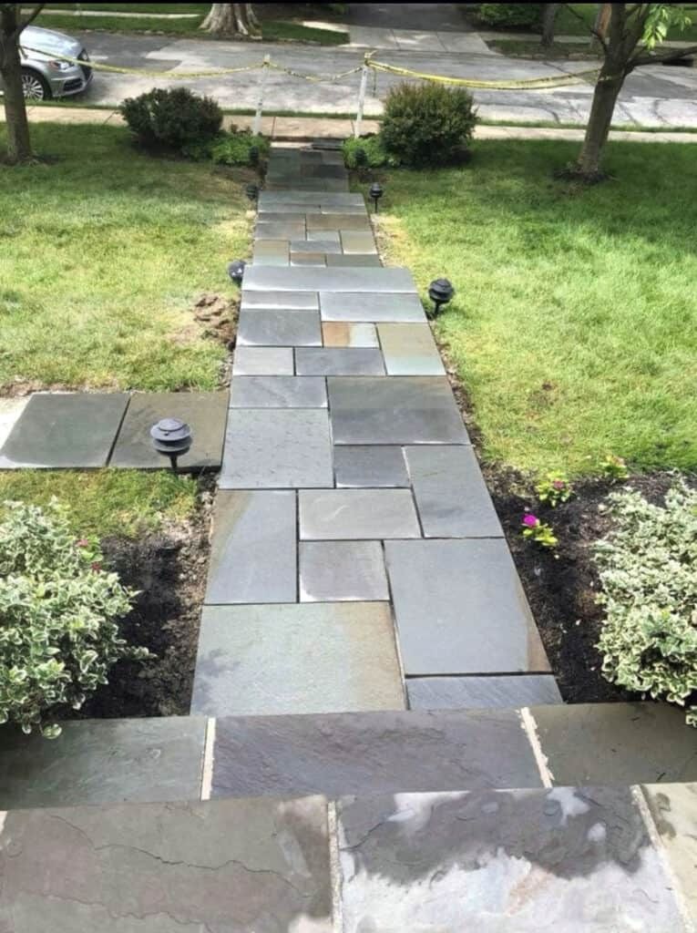 Stone walkway with steps leading towards a home, flanked by grass, bushes and low lights.